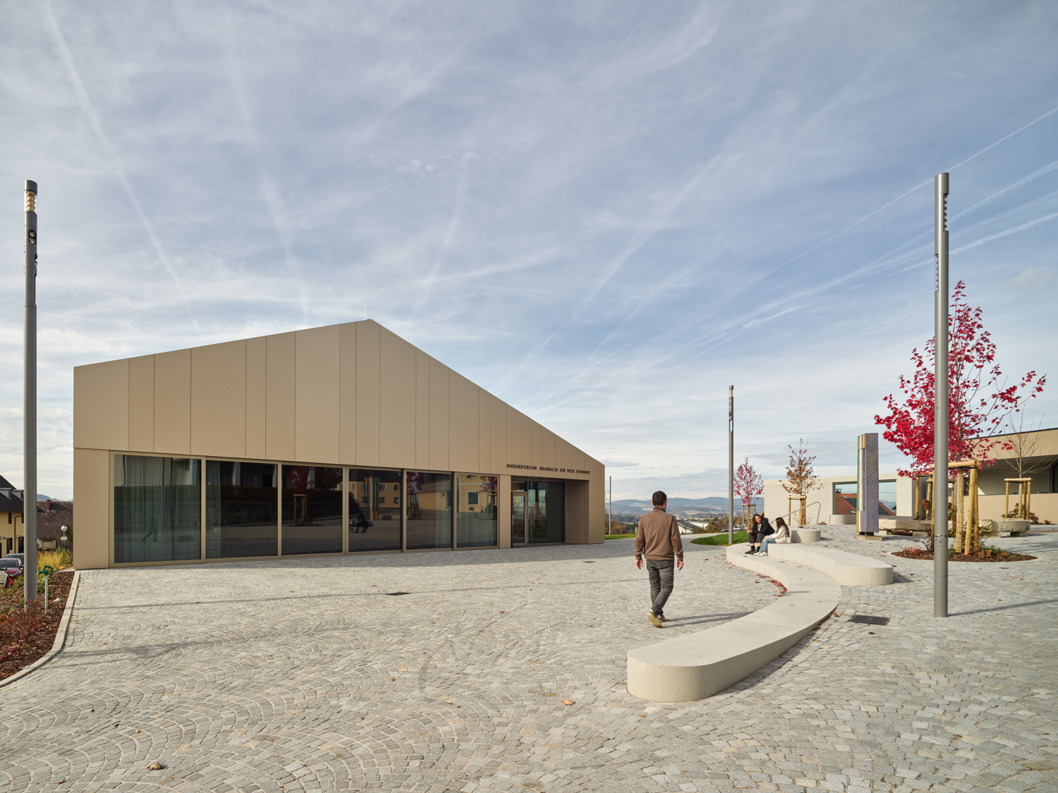 Moderner, beiger Musikverein-Gebäude mit großen Fenstern, davor gepflasterter Platz mit Sitzbänken, Bäumen mit rotem Laub und drei Menschen, eine Person geht.