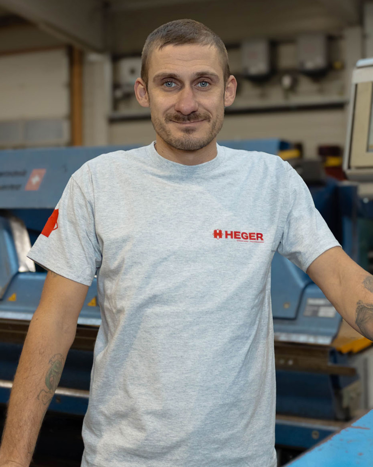 Mann mit kurzem Haar und Bart trägt ein graues T-Shirt mit rotem Heger-Logo, steht in einer Werkstatt vor einer Maschine.