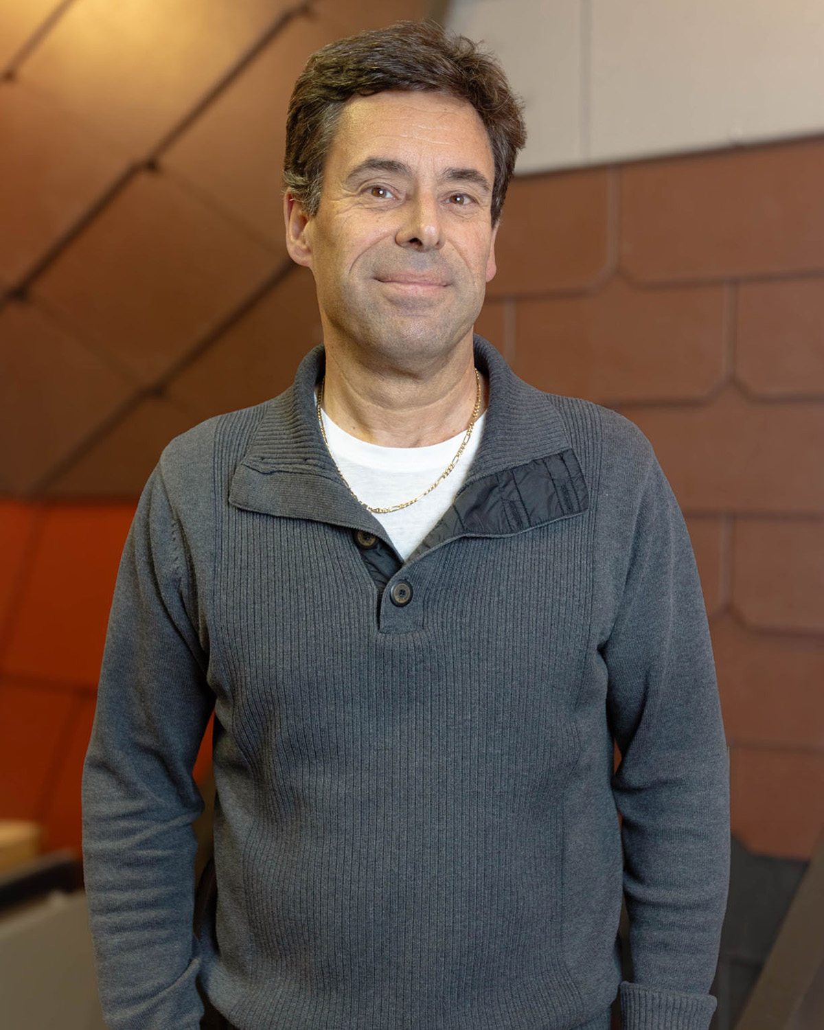 Mann mit dunklem Haar trägt grauen Pullover und goldene Halskette, lächelt leicht in die Kamera.
