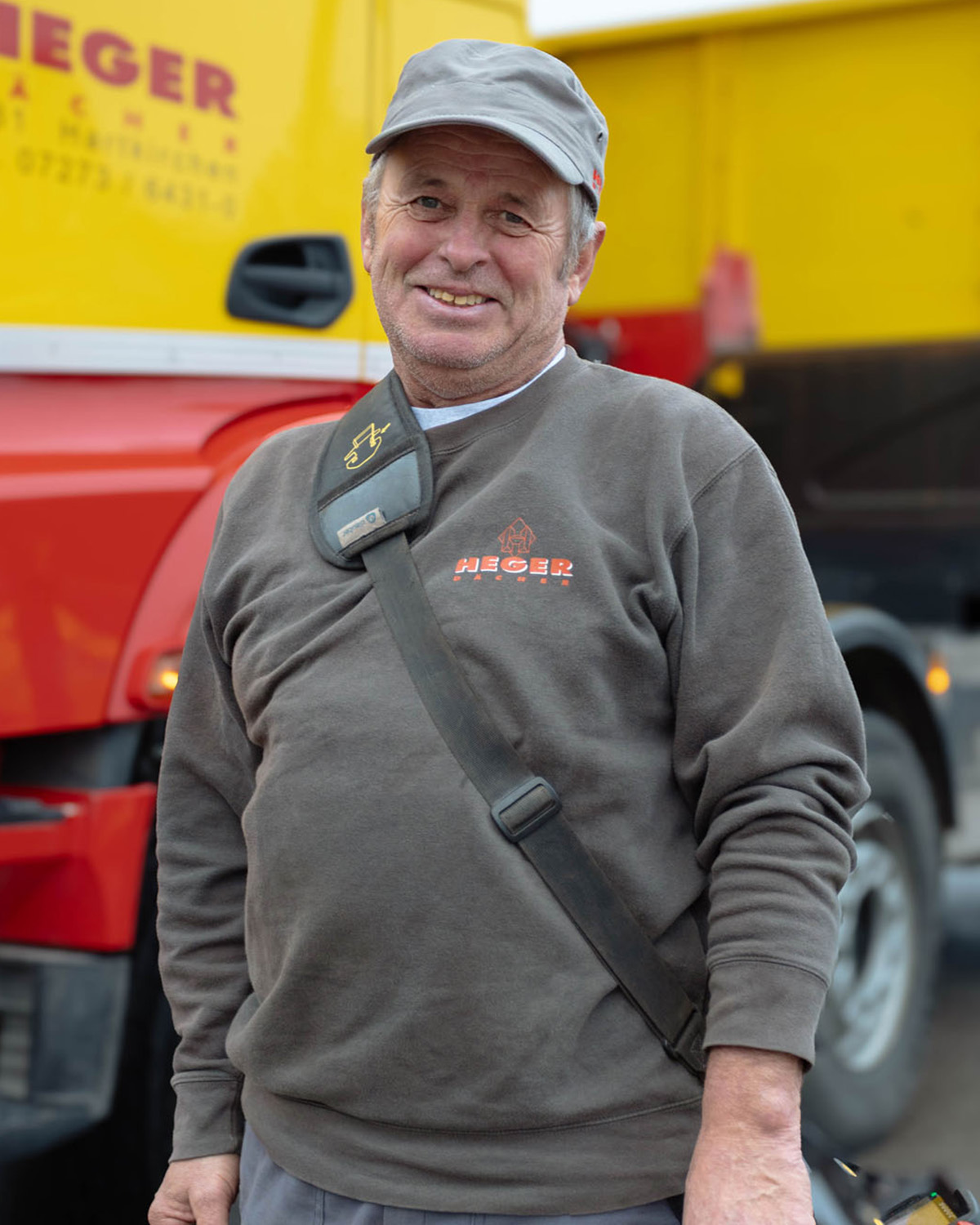 Mann mit grauem Kapperl und grauem Pullover mit Aufschrift 'HEGER' steht vor einem gelb-roten LKW.