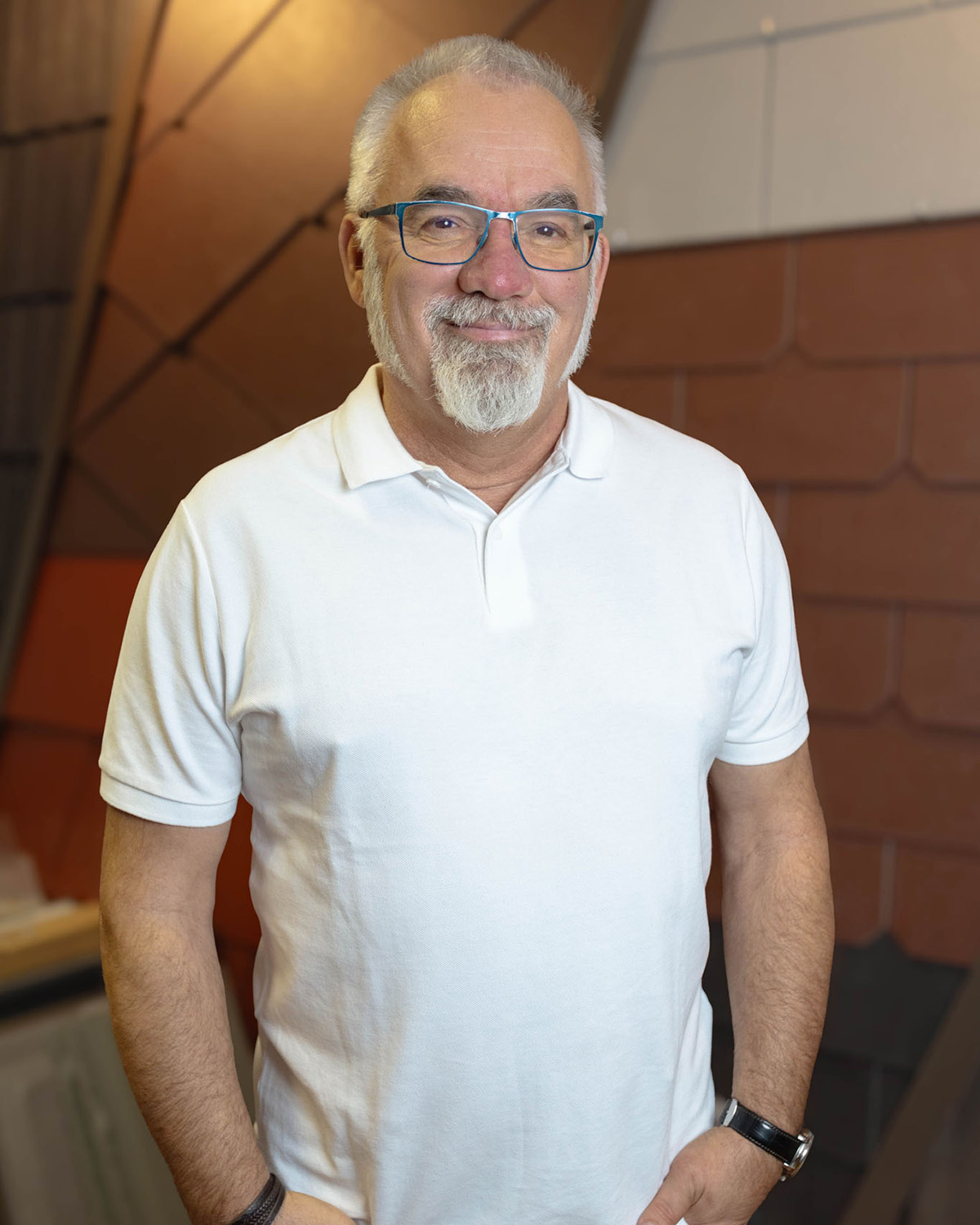 Älterer Mann mit weißem Spitzbart und blauer Brille trägt weißes Poloshirt und lächelt.