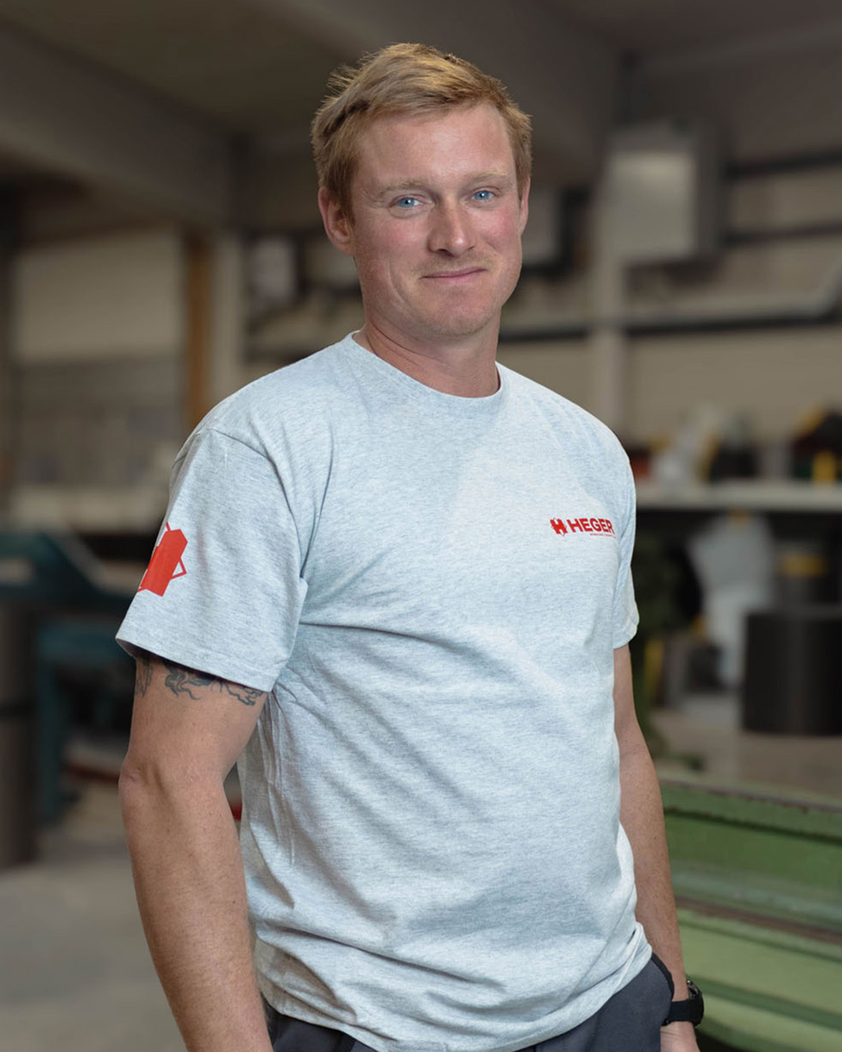 Mann mit kurzem blondem Haar trägt graues T-Shirt mit rotem Logo in einer Werkstatt.