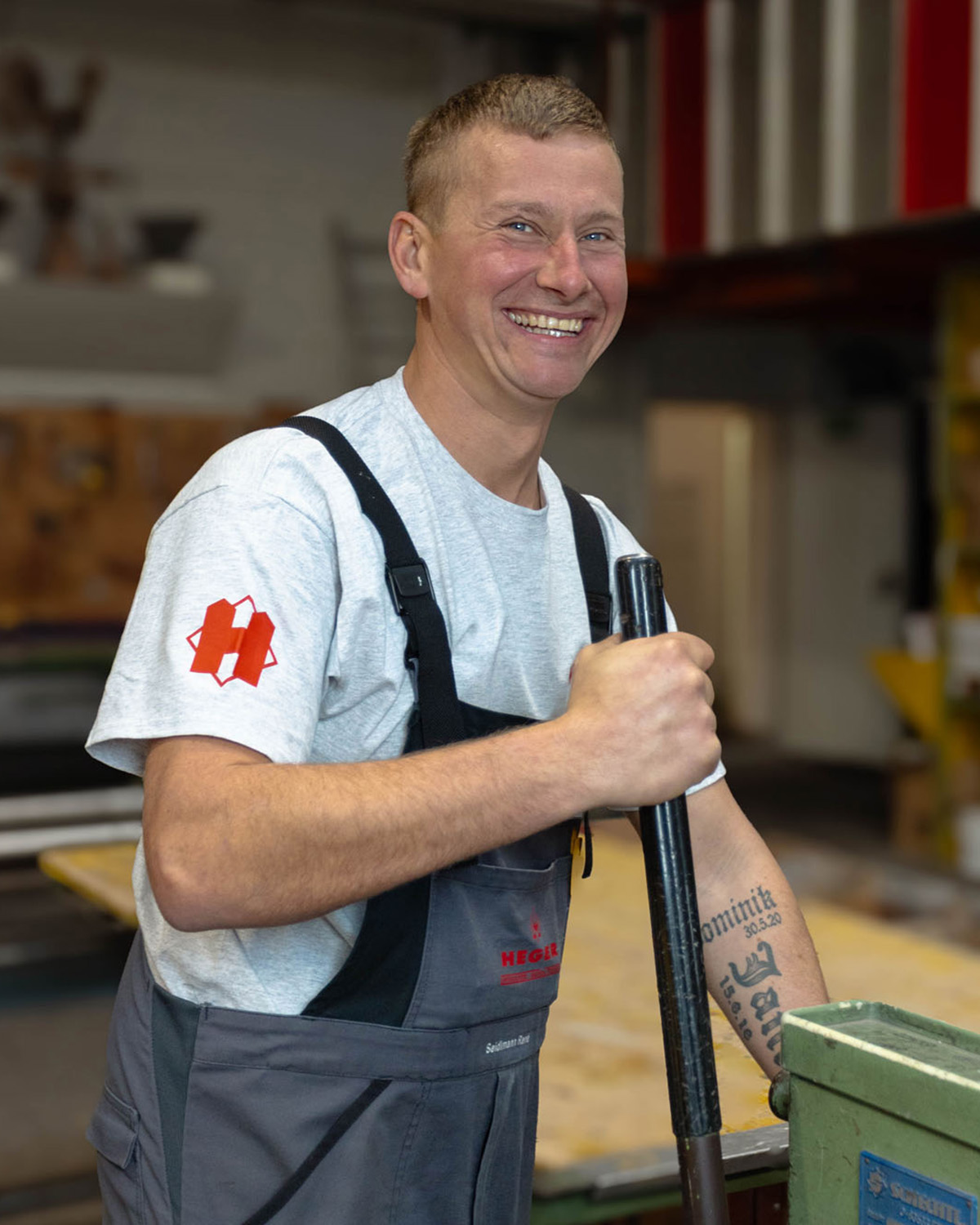 Lächelnder Mann in grauem T-Shirt und Arbeitslatzhose hält ein Werkzeug in einer Werkstatt.