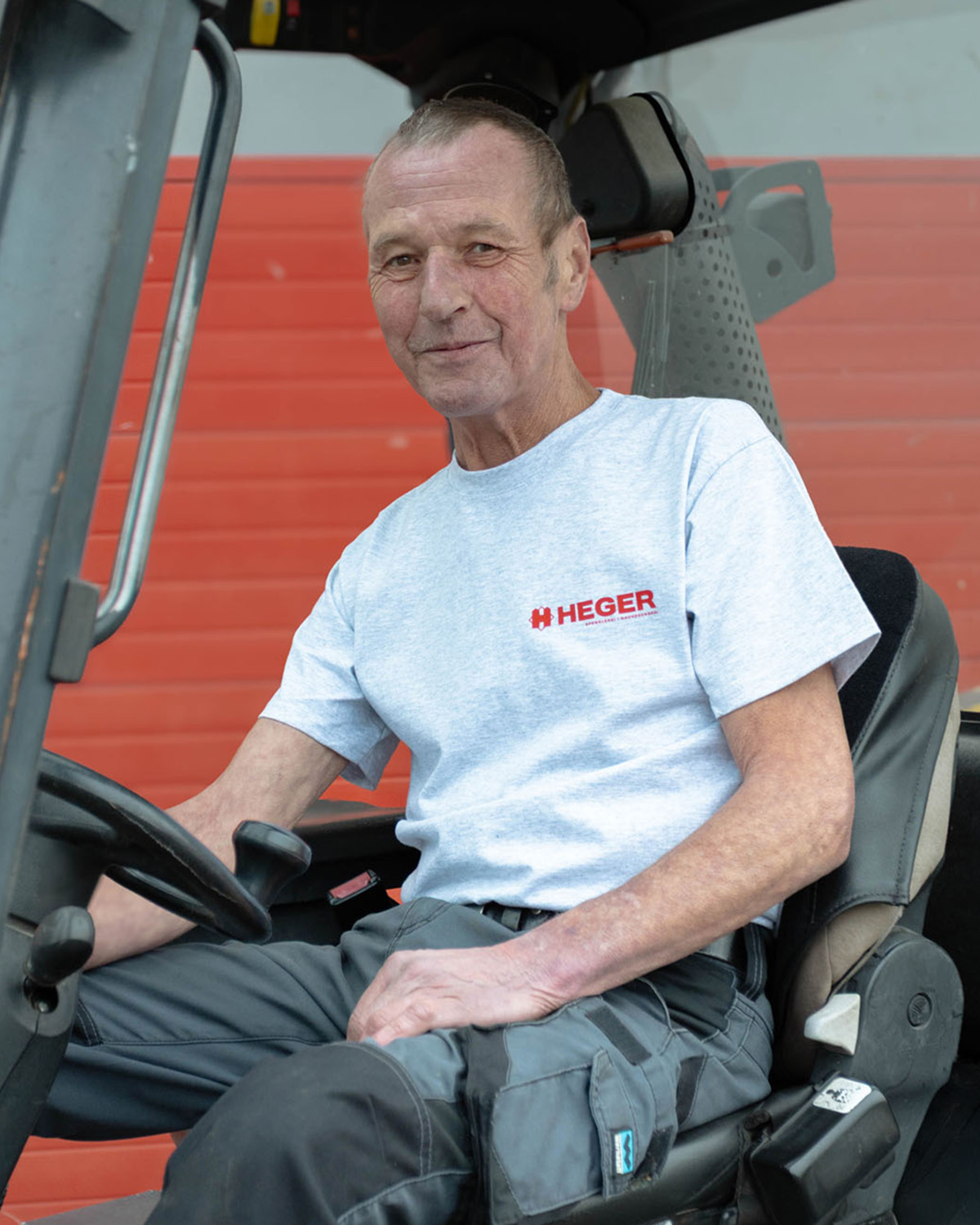 Älterer Mann mit grauem T-Shirt mit HEGER-Logo sitzt auf dem Fahrersitz eines Fahrzeugs vor einer roten Wand.