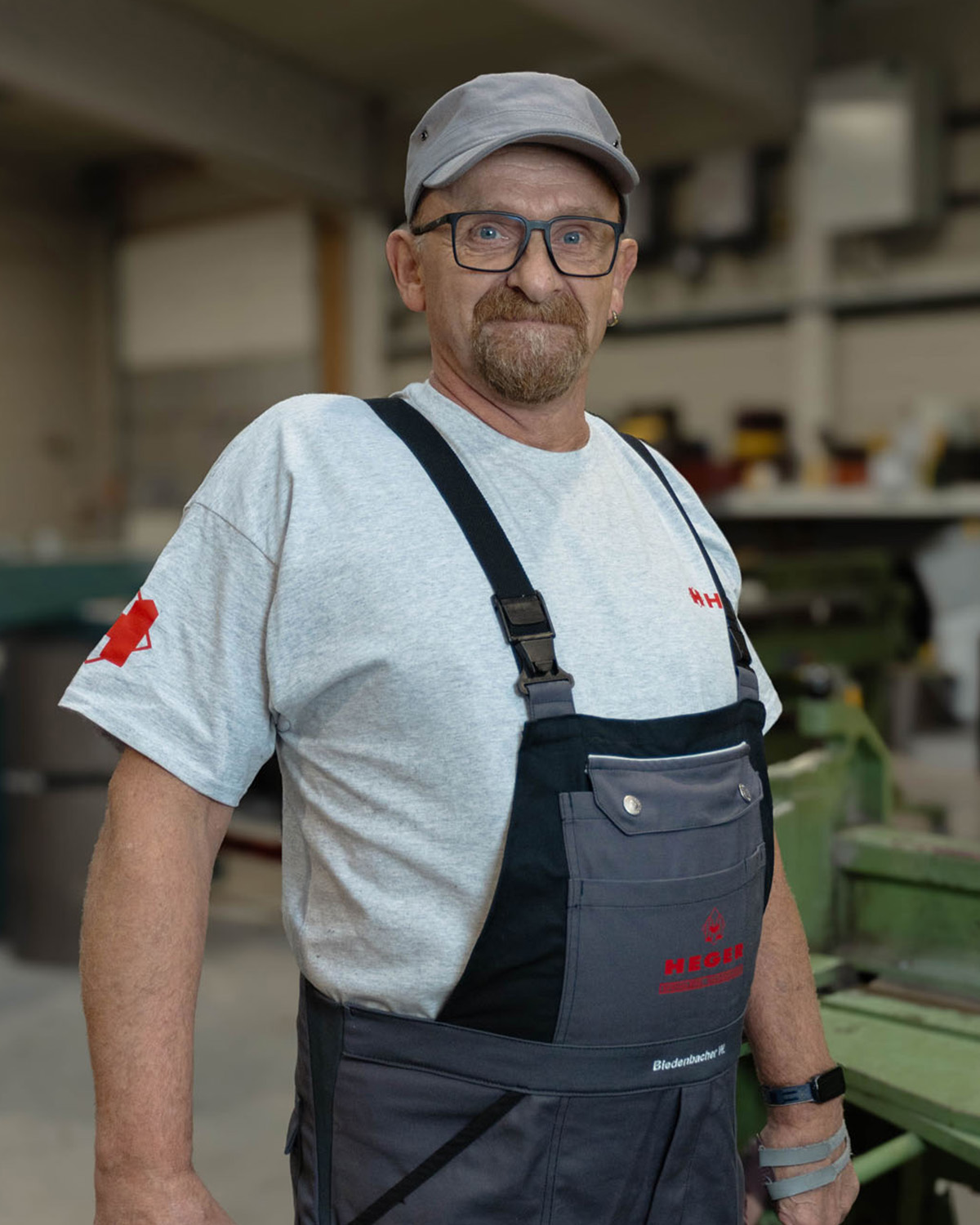 Älterer Mann mit grauem Hemd, schwarzer Latzhose, Baseballkappe und Brille in einer Werkstatt.