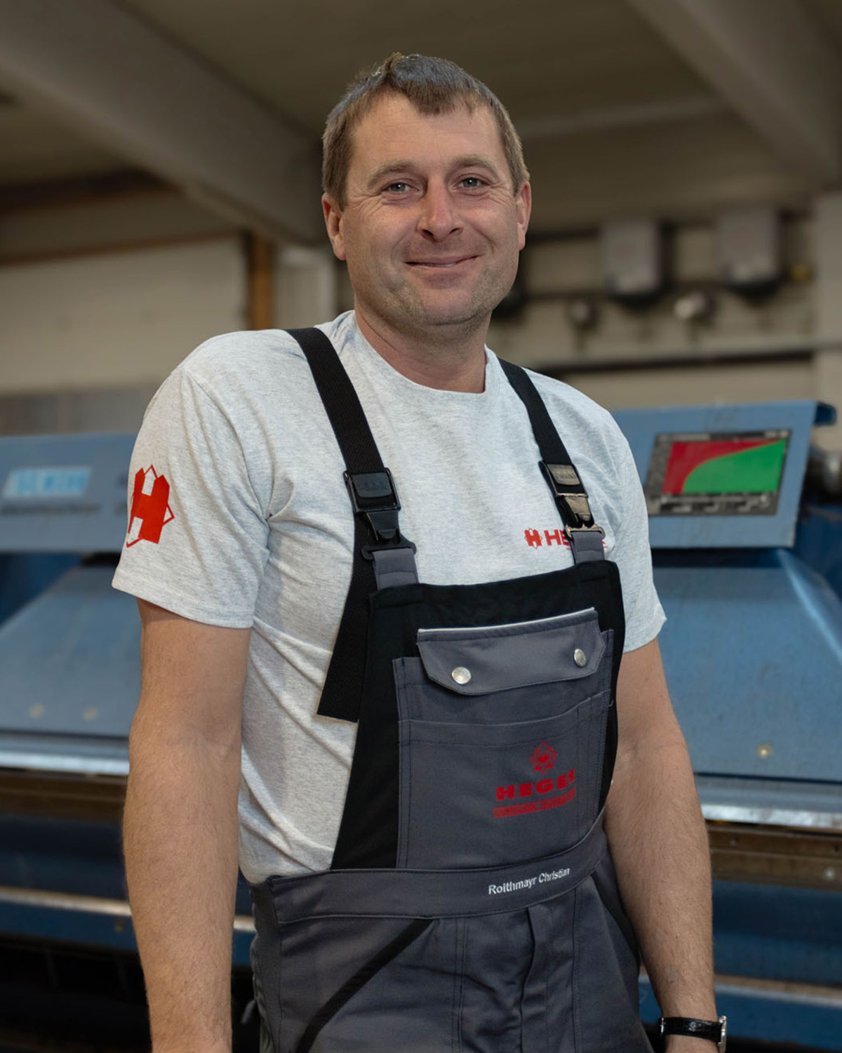 Lächelnder Mann in grauem T-Shirt und Arbeitslatzhose mit Firmenlogo vor Industriemaschine.