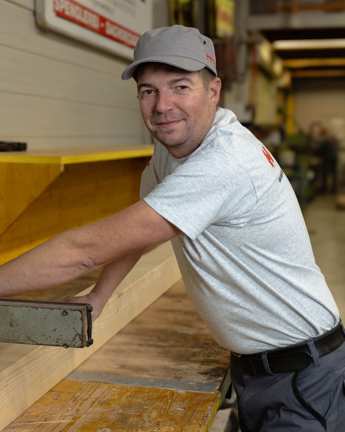 Mann mit grauem T-Shirt und grauer Kappe arbeitet an einem Holztisch in einer Werkstatt.