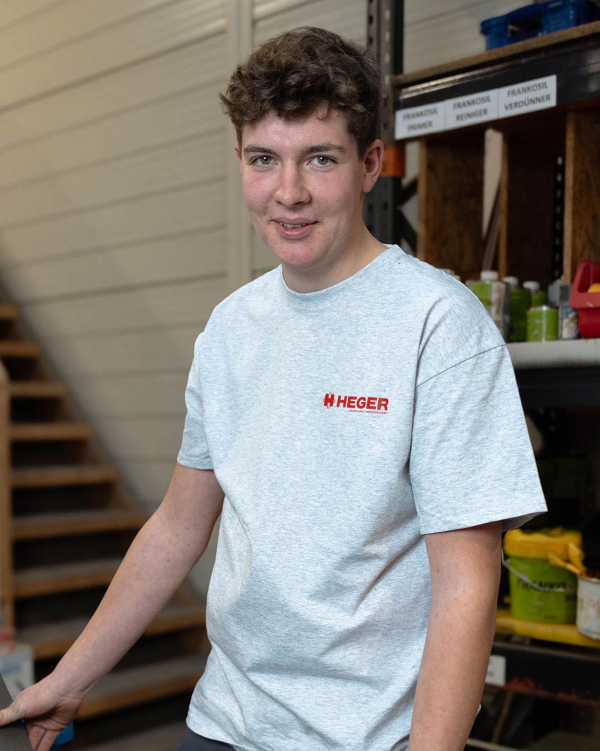 Junger Mann mit kurzem braunem Haar trägt ein hellgraues T-Shirt mit rotem Heger-Logo und steht in einer Werkstatt neben einem Regal.