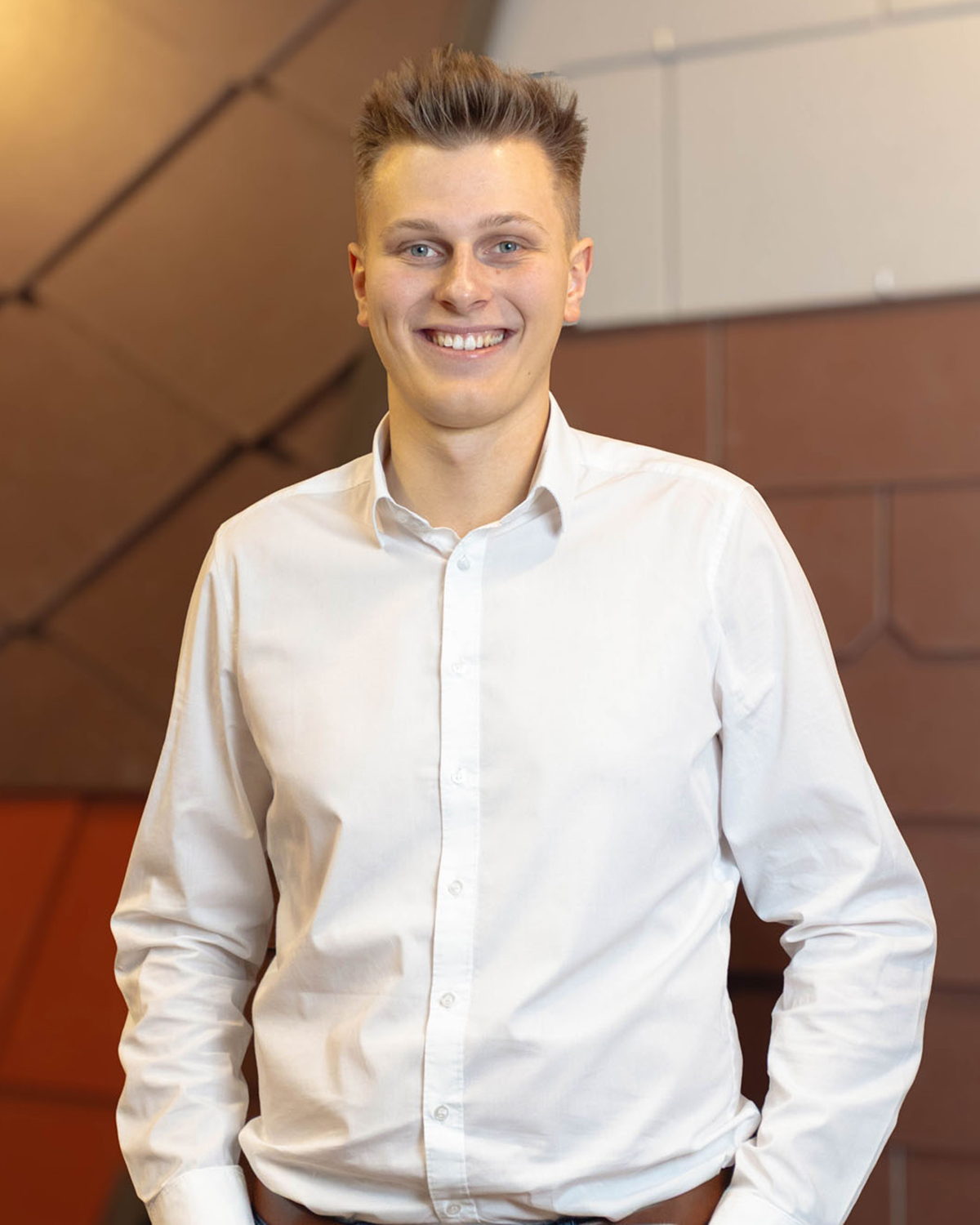 Junger Mann mit kurzen blonden Haaren lächelt und trägt ein weißes Hemd vor einer braunen Wand.