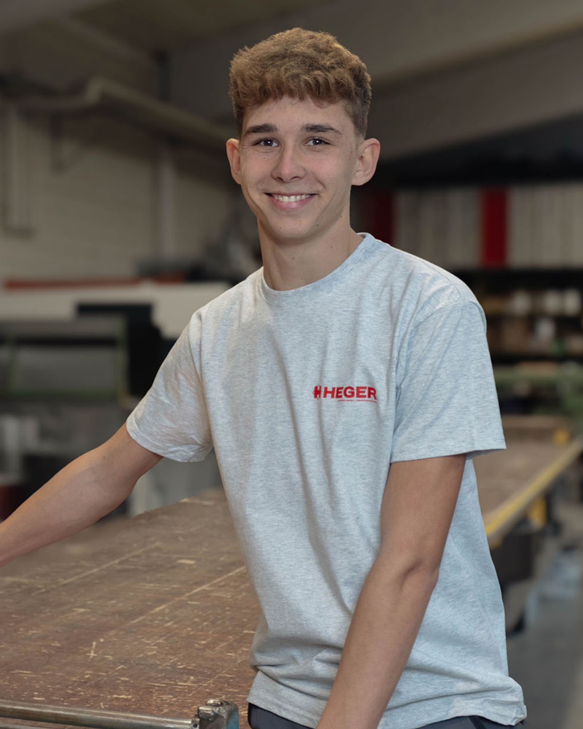 Junger Mann mit kurzem lockigem Haar lächelt und trägt ein graues T-Shirt mit dem Logo HEGER, steht in einer Werkstatt.