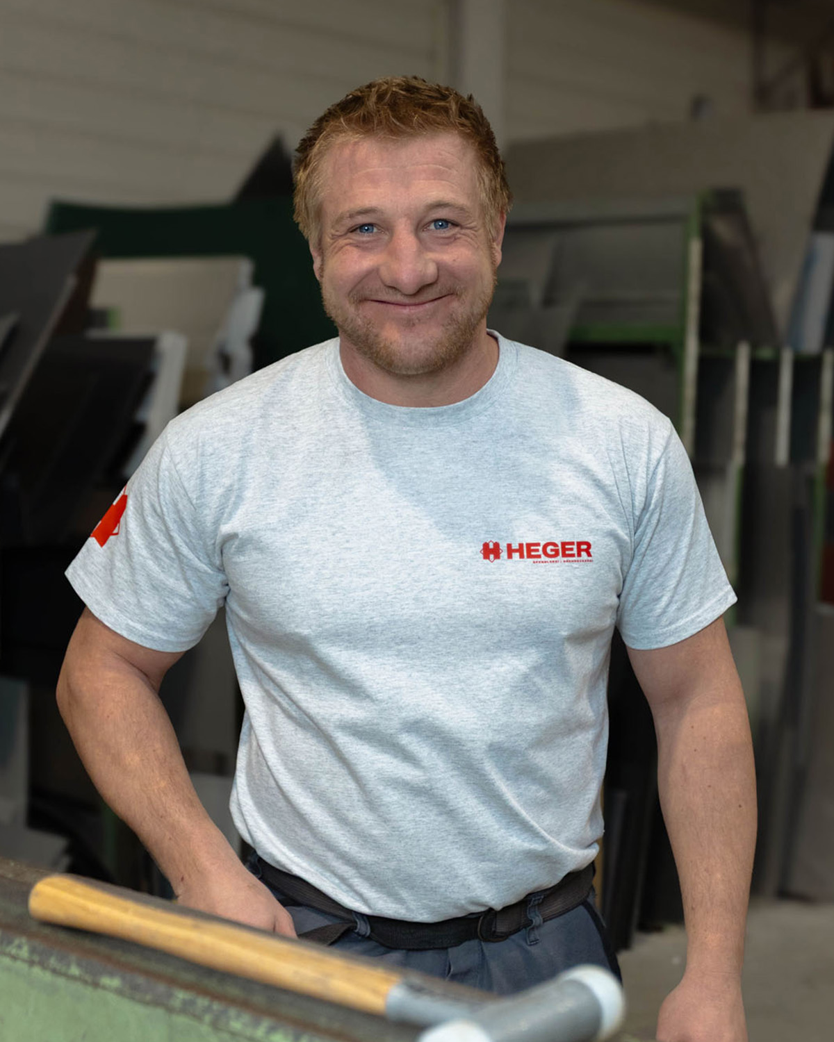 Lächelnder Mann mit kurzen blonden Haaren trägt ein graues Hemd mit rotem HEGER-Logo in einer Werkstatt.