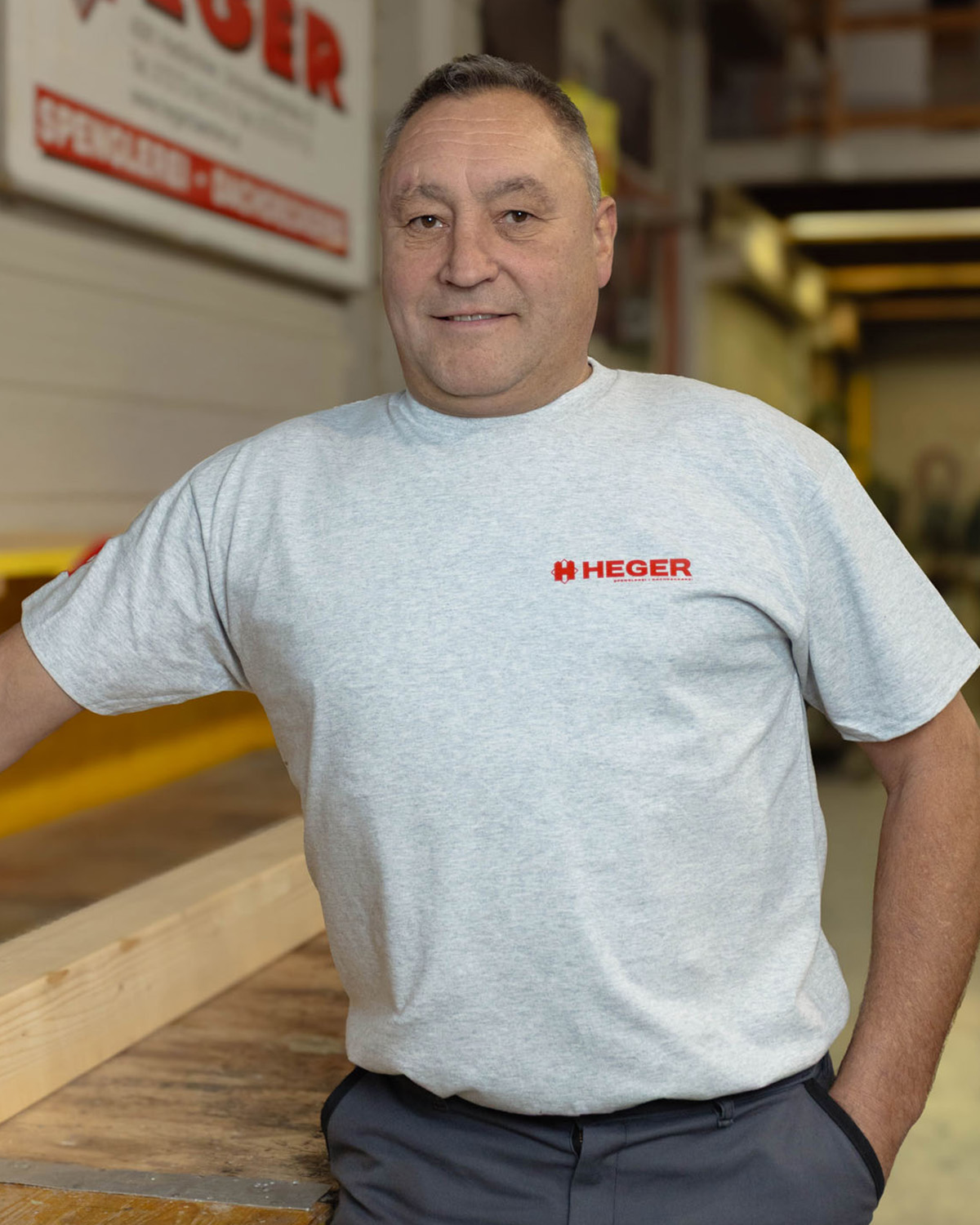 Mann in grauem T-Shirt mit rotem HEGER-Logo lehnt in einer Werkstatt an einem Holztisch.