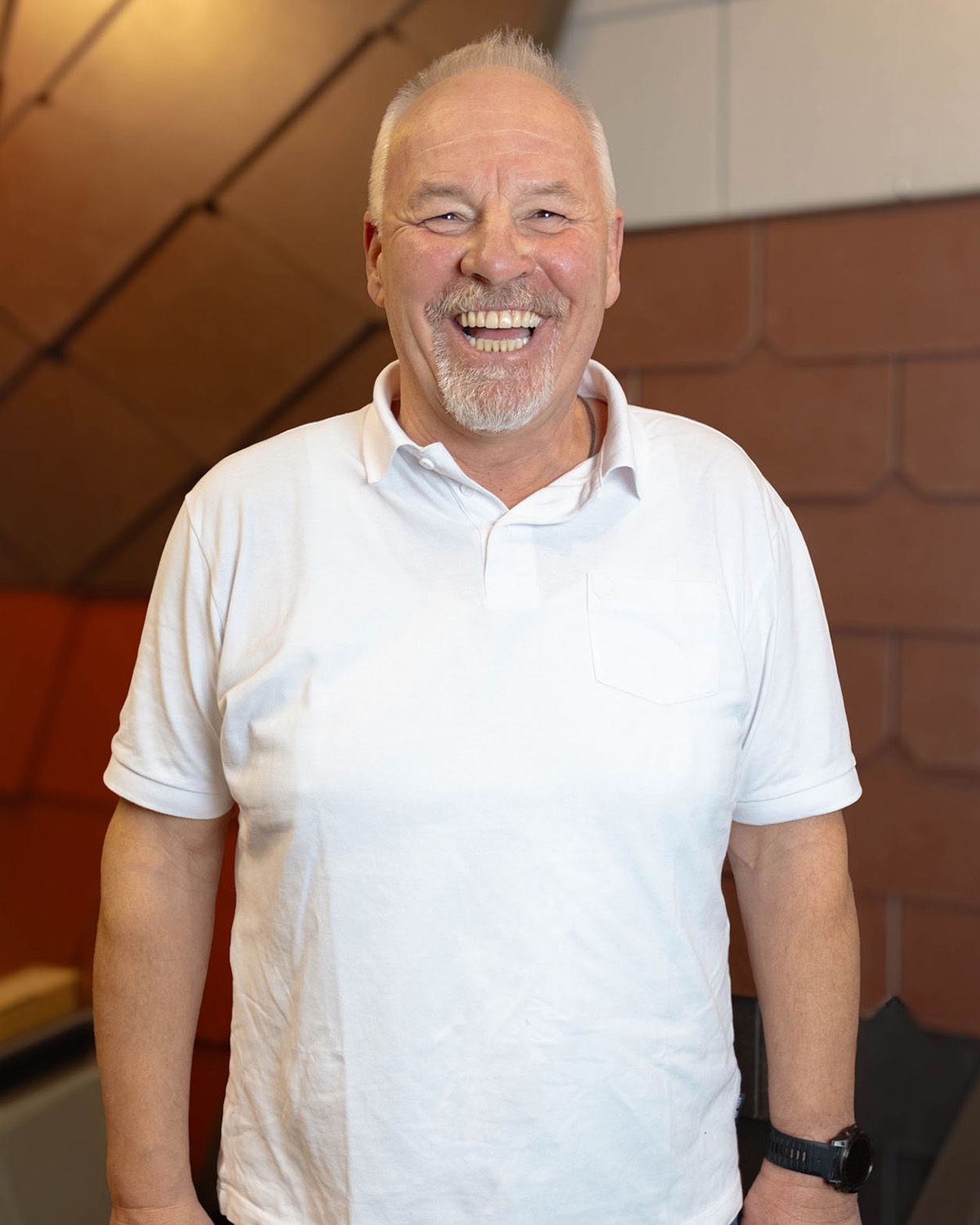 Älterer Mann mit weißem kurzärmligem Poloshirt und schwarzer Armbanduhr lächelt freundlich in die Kamera.