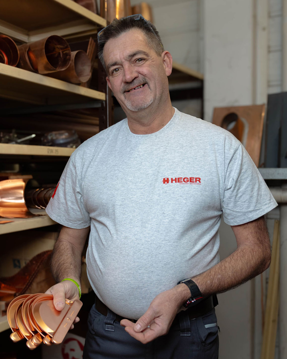 Männlicher Handwerker in grauem T-Shirt mit Heger-Logo hält mehrere kupferfarbene Metallteile in einer Werkstatt.