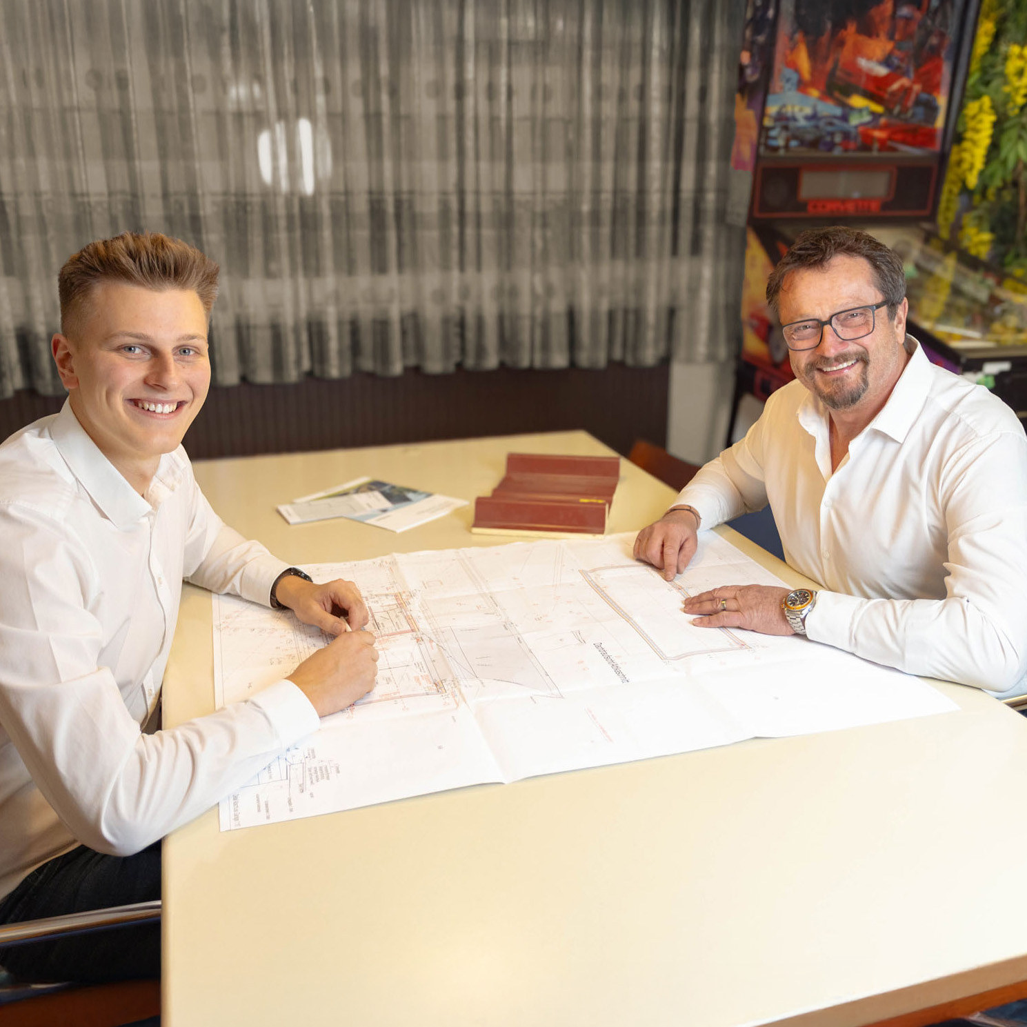 Zwei lächelnde Männer in weißen Hemden sitzen an einem Tisch und betrachten Baupläne.
