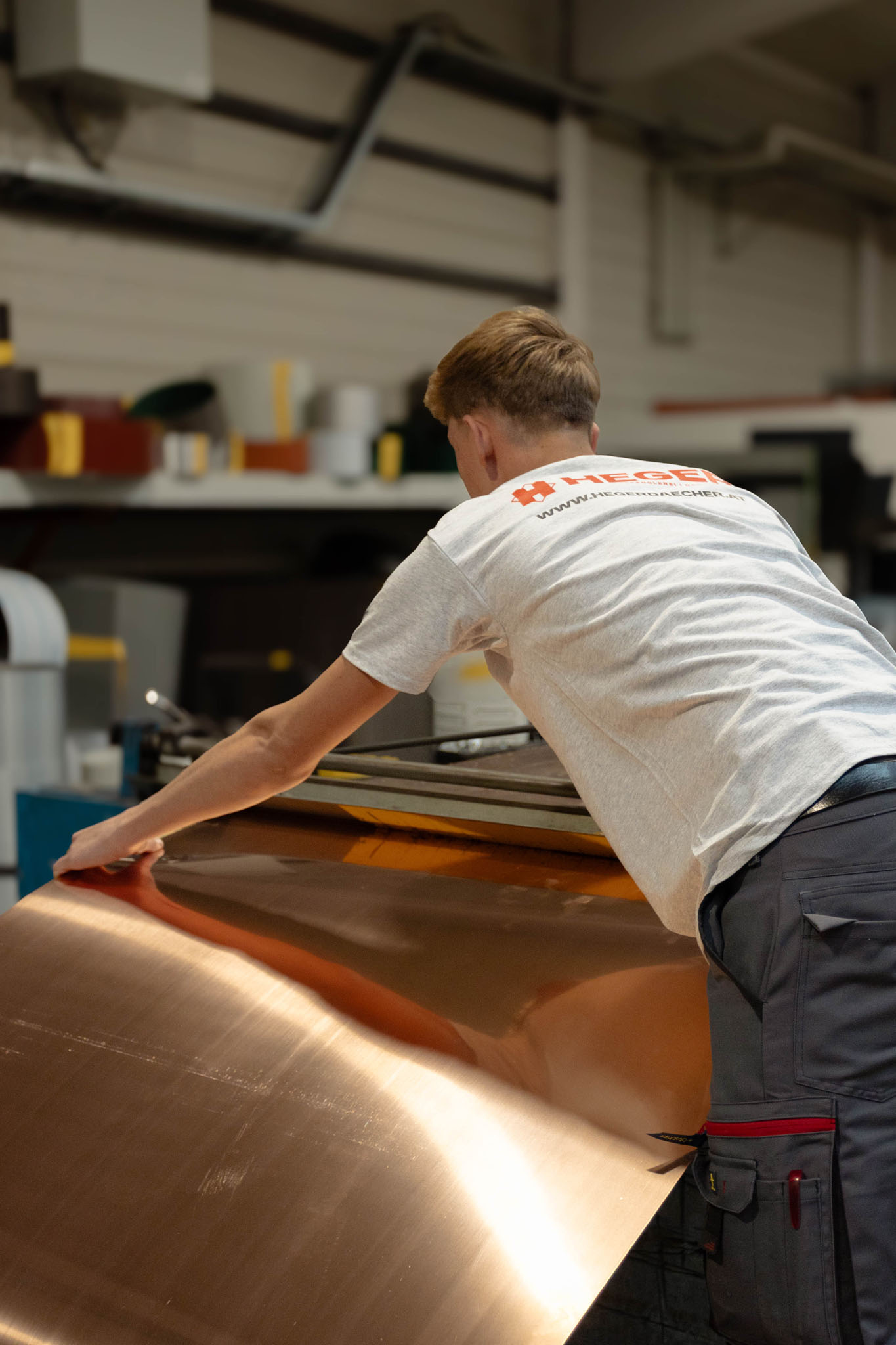 Ein Mann in einem grauen T-Shirt bearbeitet eine große glänzende Kupferplatte in einer Werkstatt.