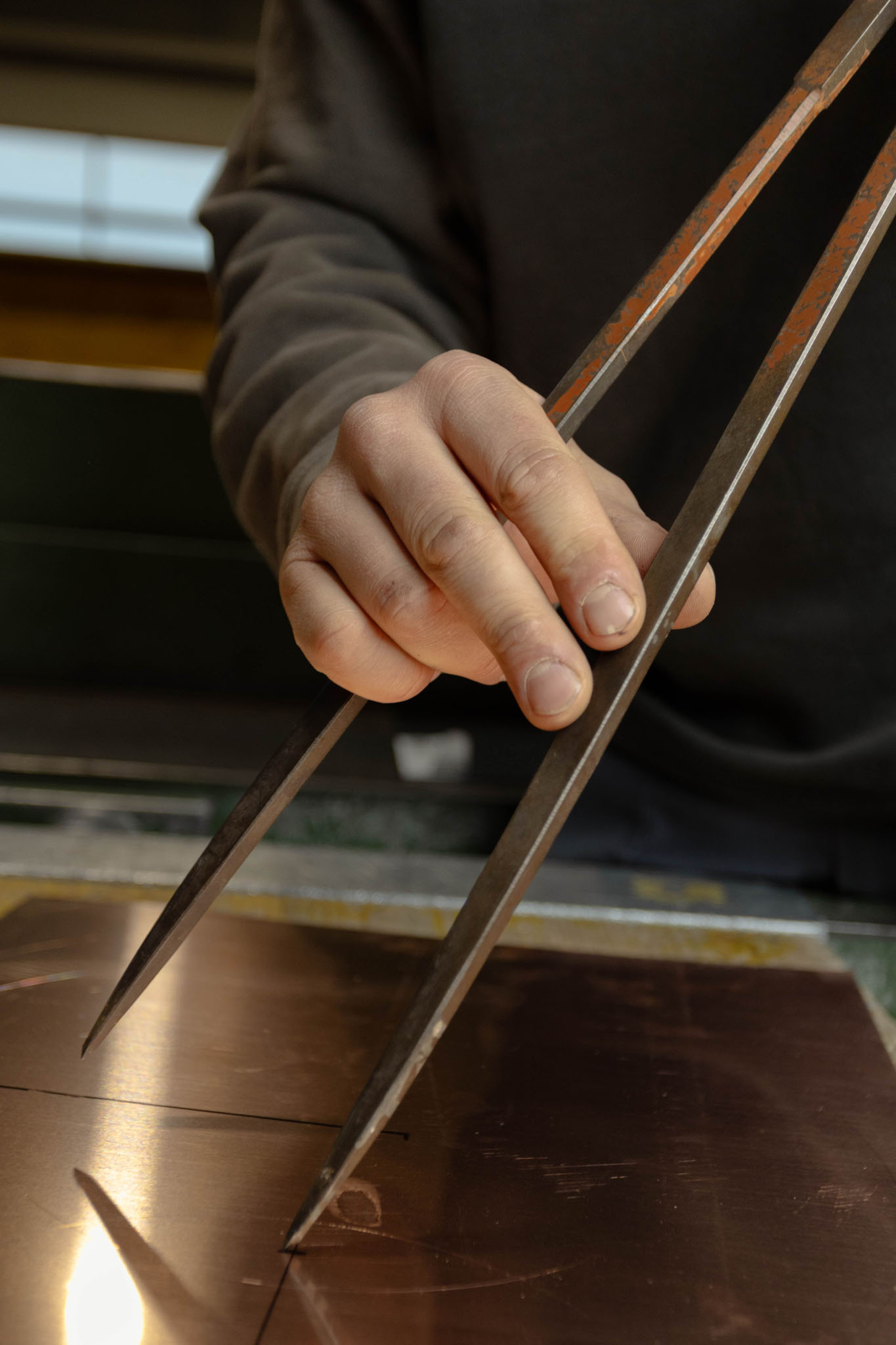 Hand hält einen großen Stiftzirkel über ein Kupferblech zum Abmessen oder Zeichnen.