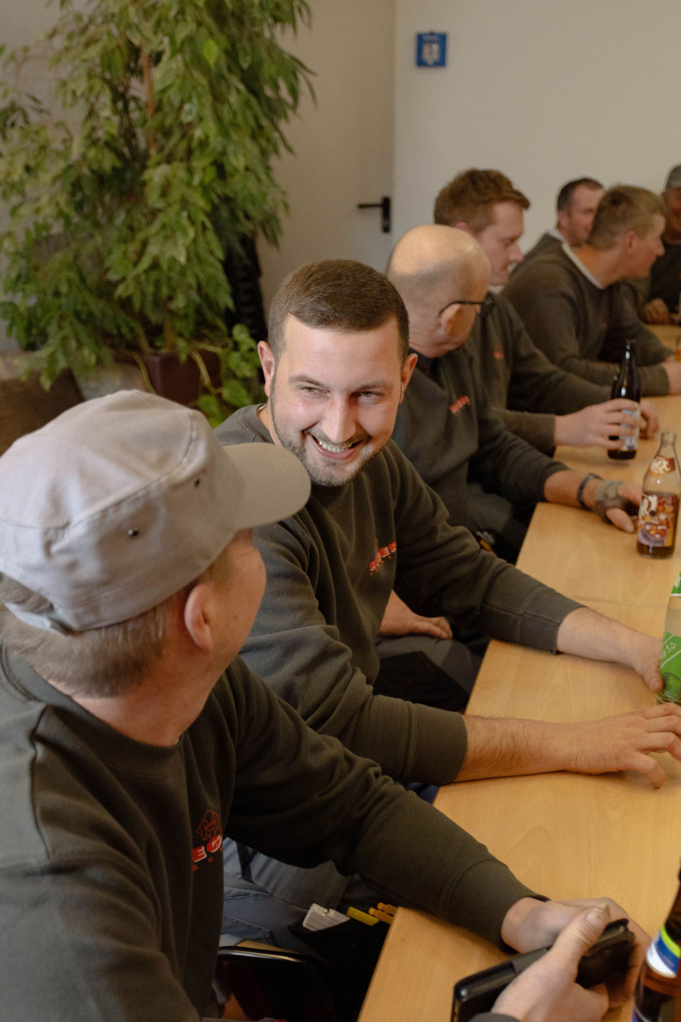 Eine Gruppe Männer in dunklen Pullovern sitzt um einen Holztisch und unterhält sich, einer lächelt besonders mit einem Mann, der eine graue Mütze trägt.