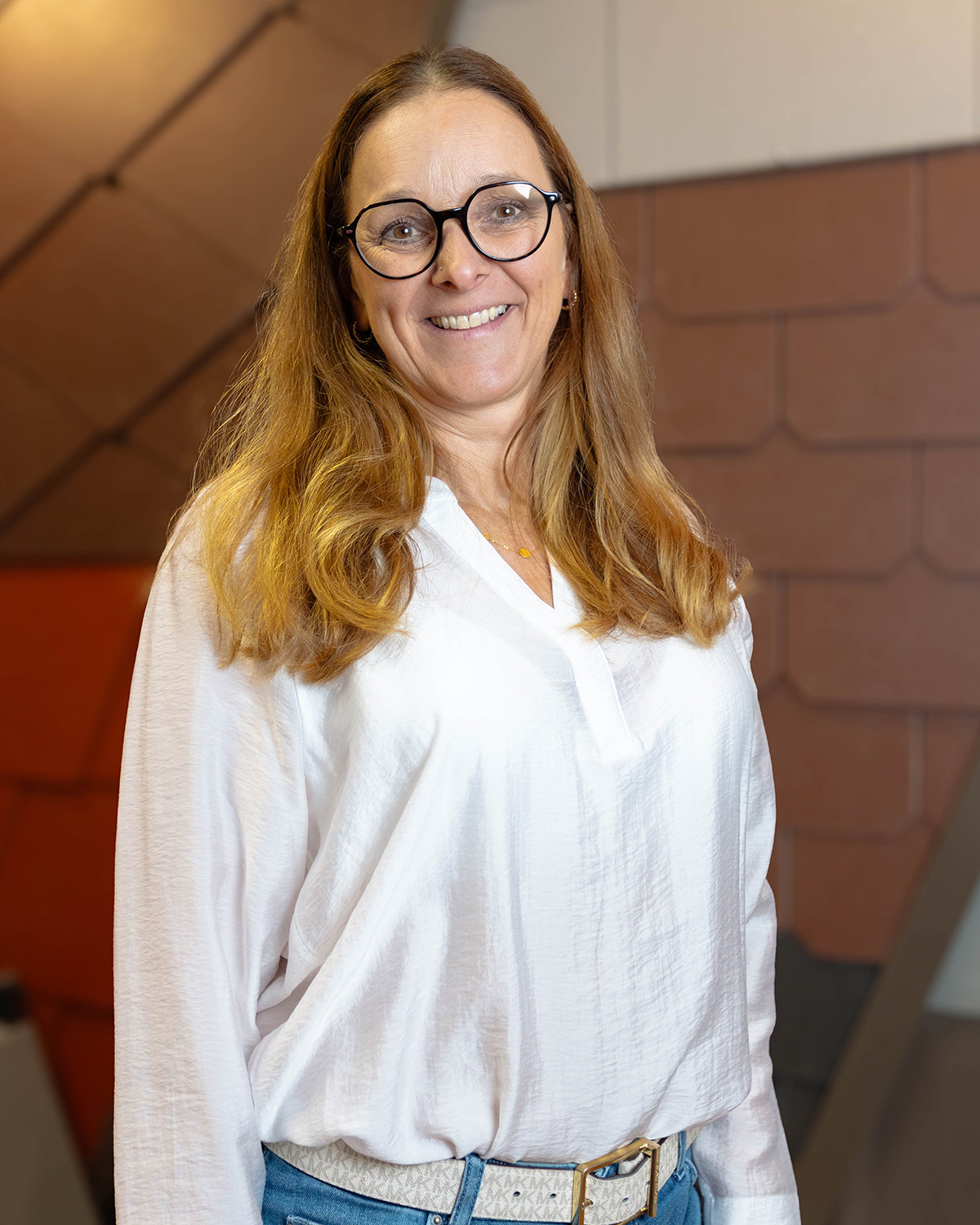 Lächelnde Frau mit langen blonden Haaren, schwarzer Brille, weißer Bluse und Jeans vor orange-braunem Hintergrund.