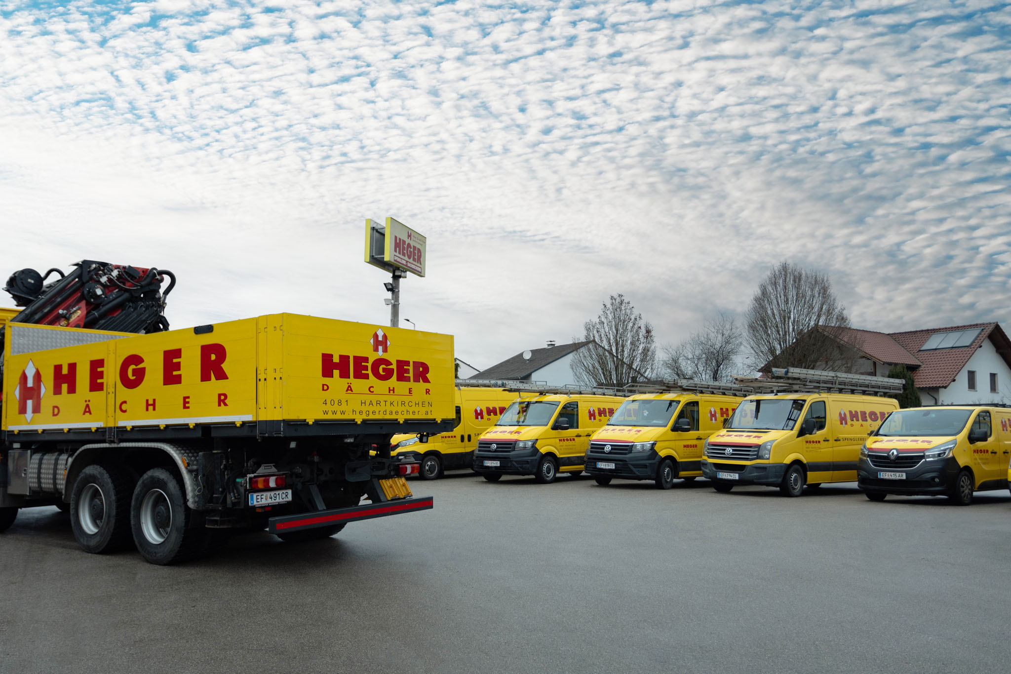 Gelbe Fahrzeuge mit der Aufschrift HEGER DÄCHER, darunter ein Lastwagen und mehrere Transporter vor einem Gebäude unter bewölktem Himmel.