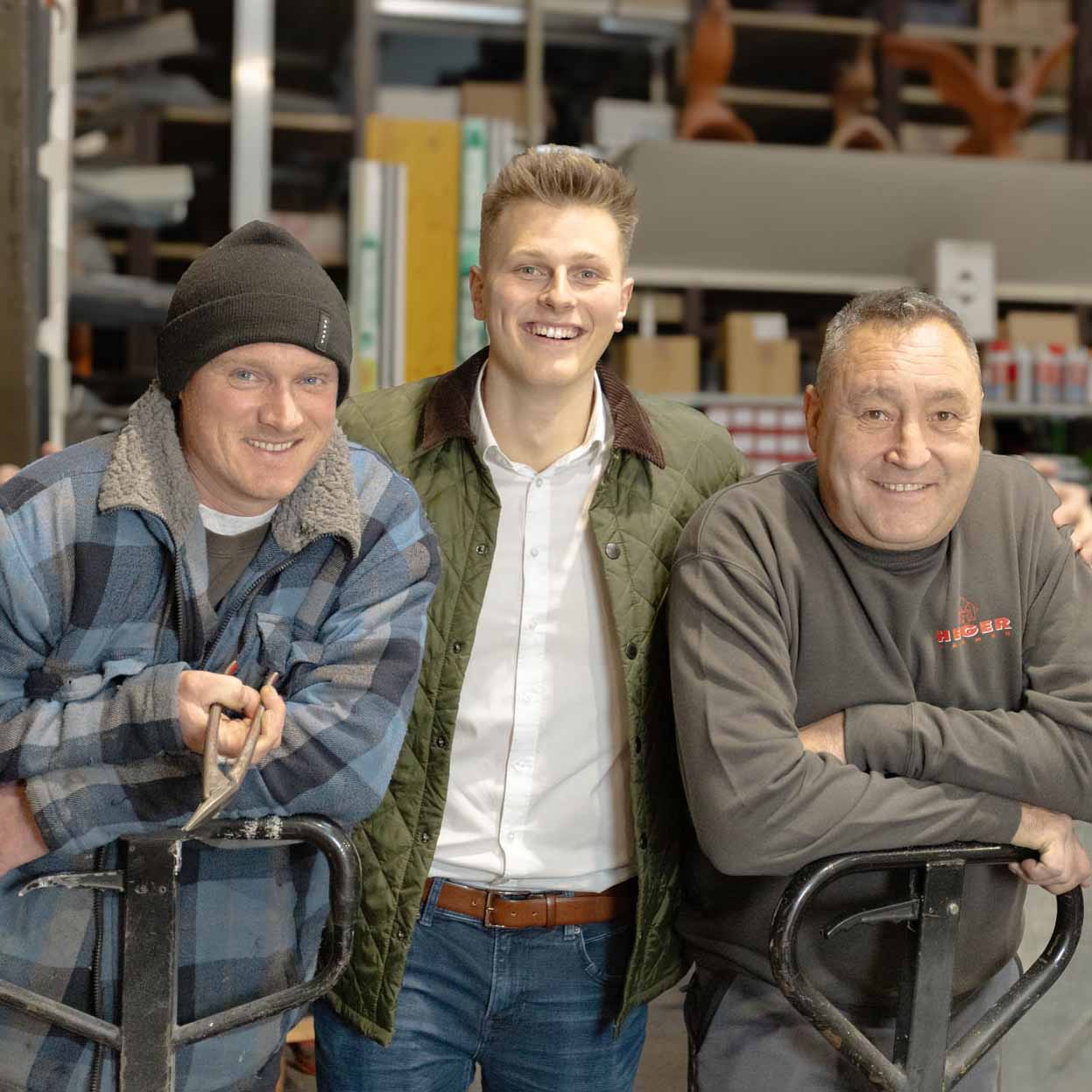 Drei lächelnde Männer mittleren Alters stehen in einem Lager, zwei lehnen an Hubwagen.