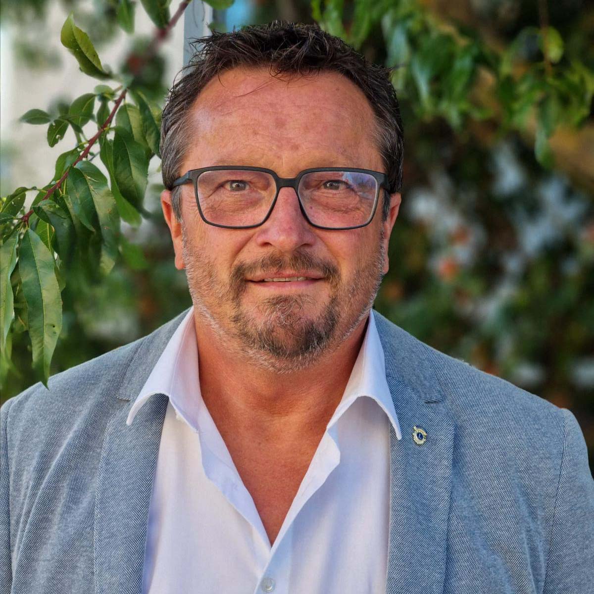 Porträt eines Mannes mit Brille, kurzen dunklen Haaren und Bart, der einen grauen Mantel und weißes Hemd trägt, vor grünen Blättern stehend.