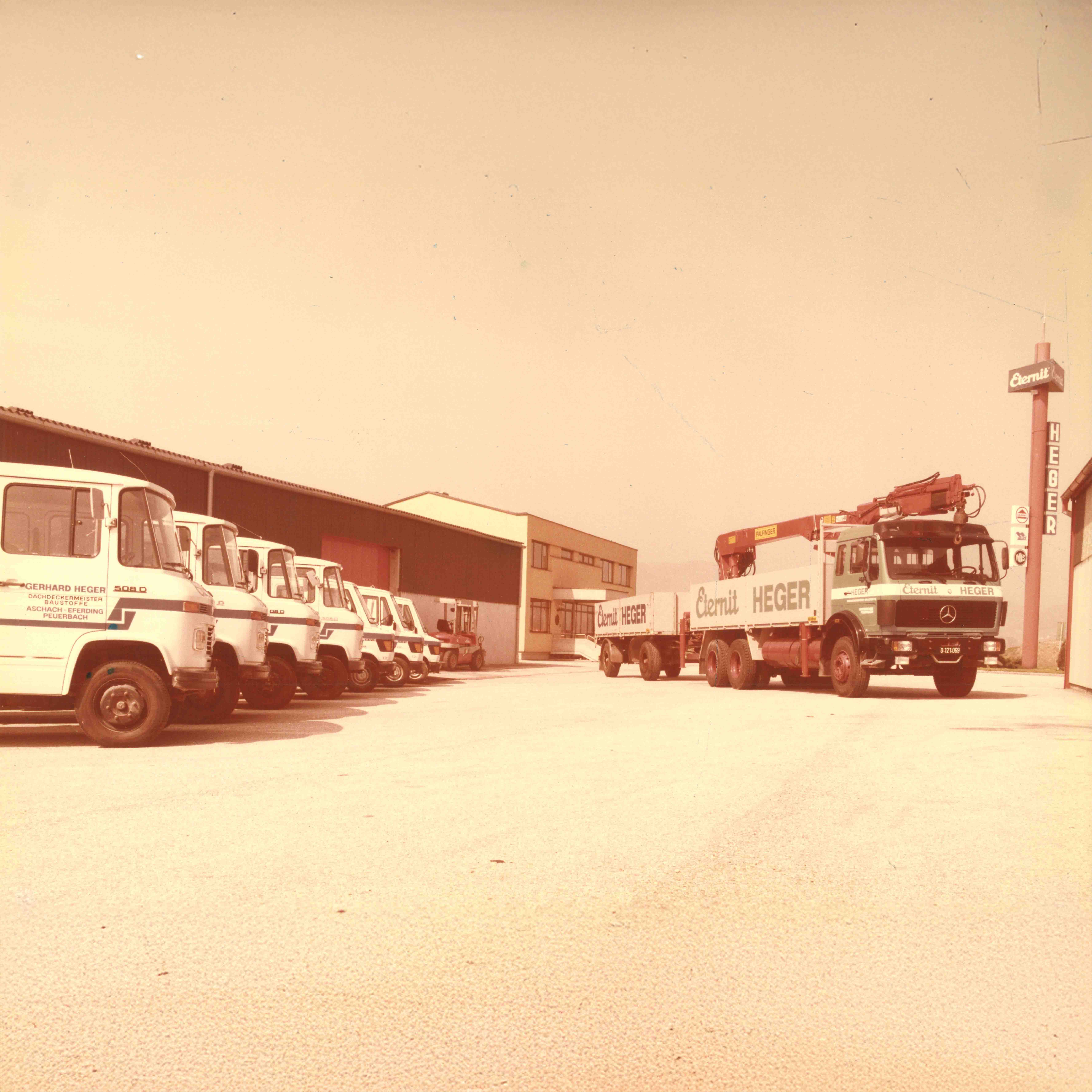 Reihe von weißen Firmen-Lkw der Firma Gerhard Heger und zwei Lkw mit Anhänger vor Lagergebäuden.