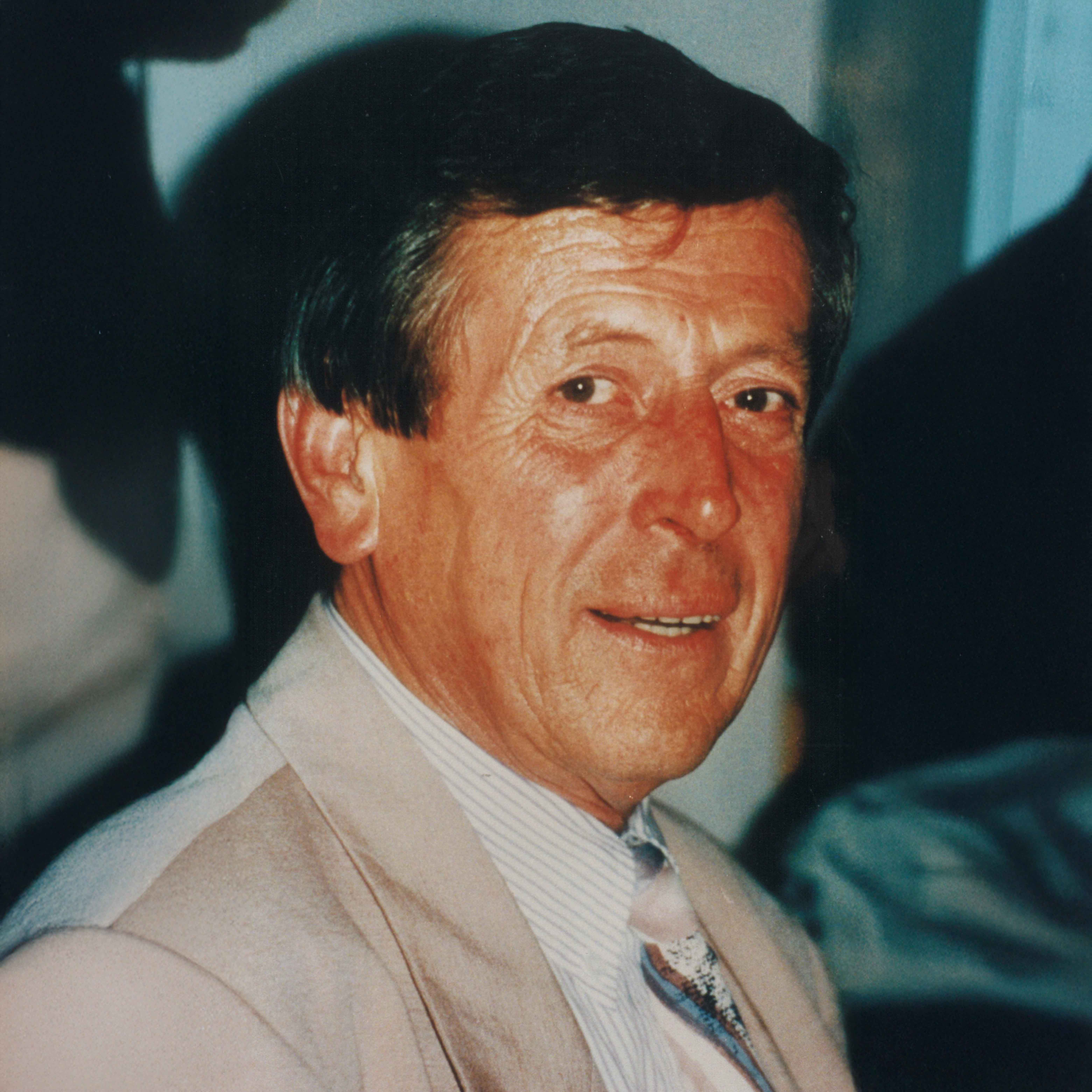 Porträt eines älteren Mannes mit kurzem dunklem Haar, der einen beigen Anzug und eine gemusterte Krawatte trägt.