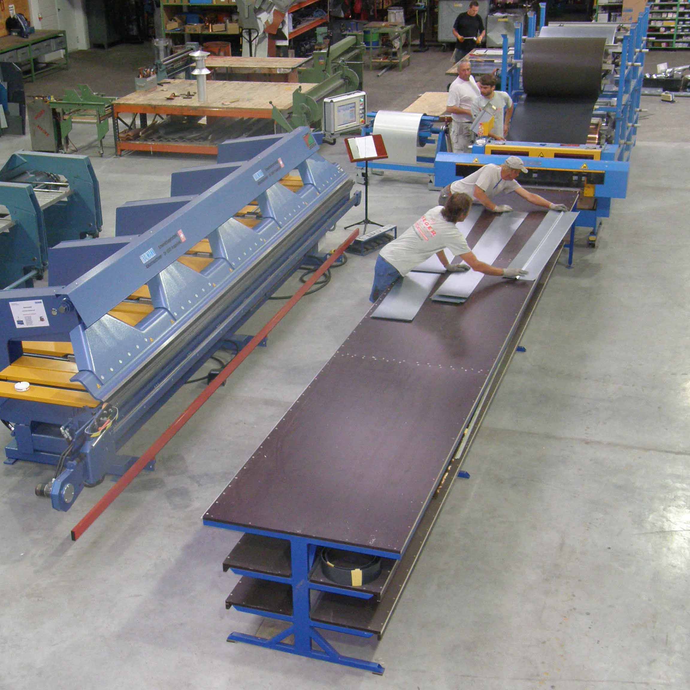 Fabrikhalle mit vier Arbeitern, die Metallbleche auf einer langen Werkbank bearbeiten und Maschinen im Hintergrund.