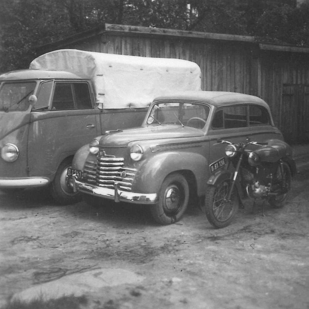 Schwarz-weiß-Foto von einem alten Lastwagen mit Plane, einem klassischen Auto und einem Motorrad, die vor einer Holzhütte geparkt sind.