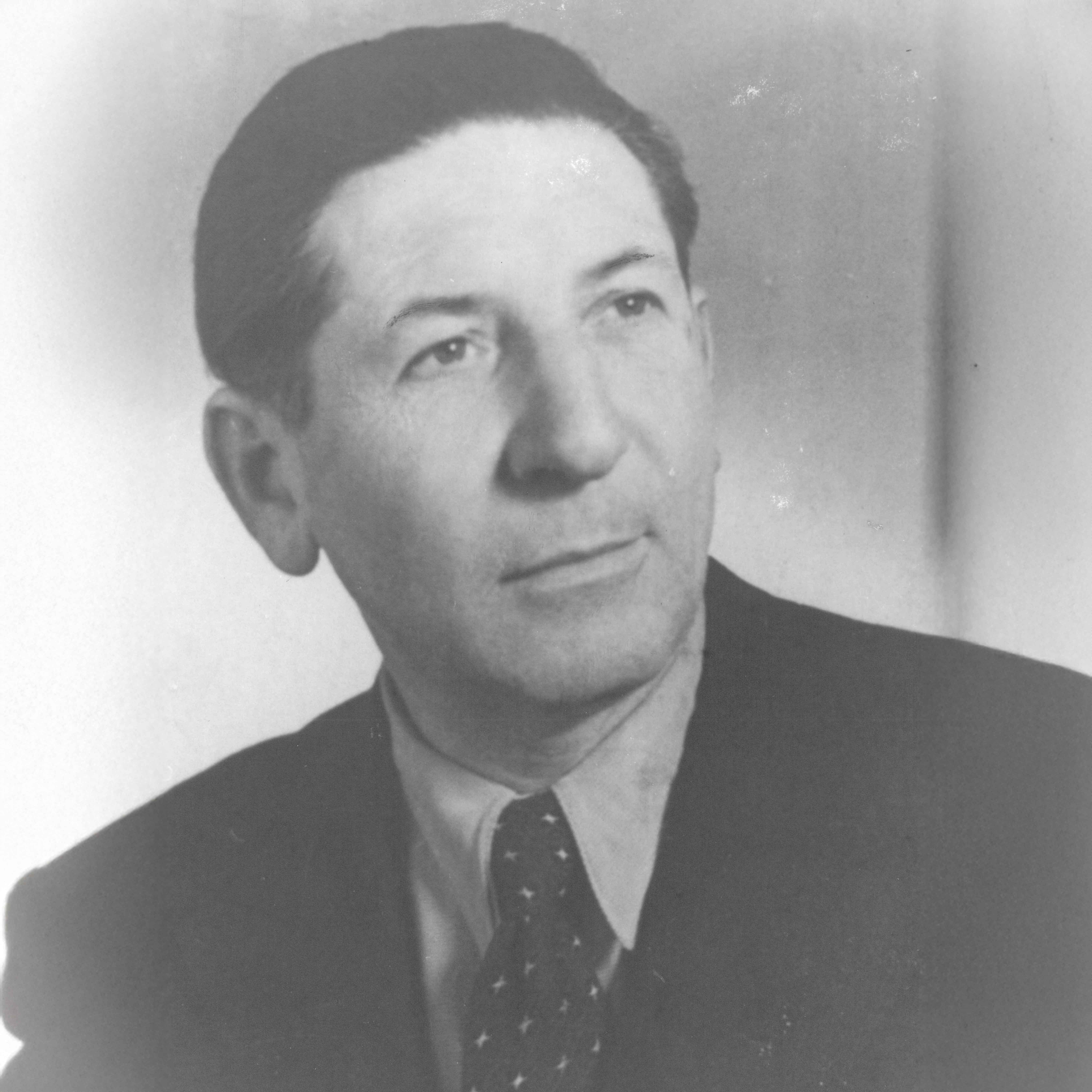 Schwarzweiß-Porträt eines Mannes mit zurückgekämmtem Haar, Anzug und gemusterter Krawatte, der nach oben rechts blickt.