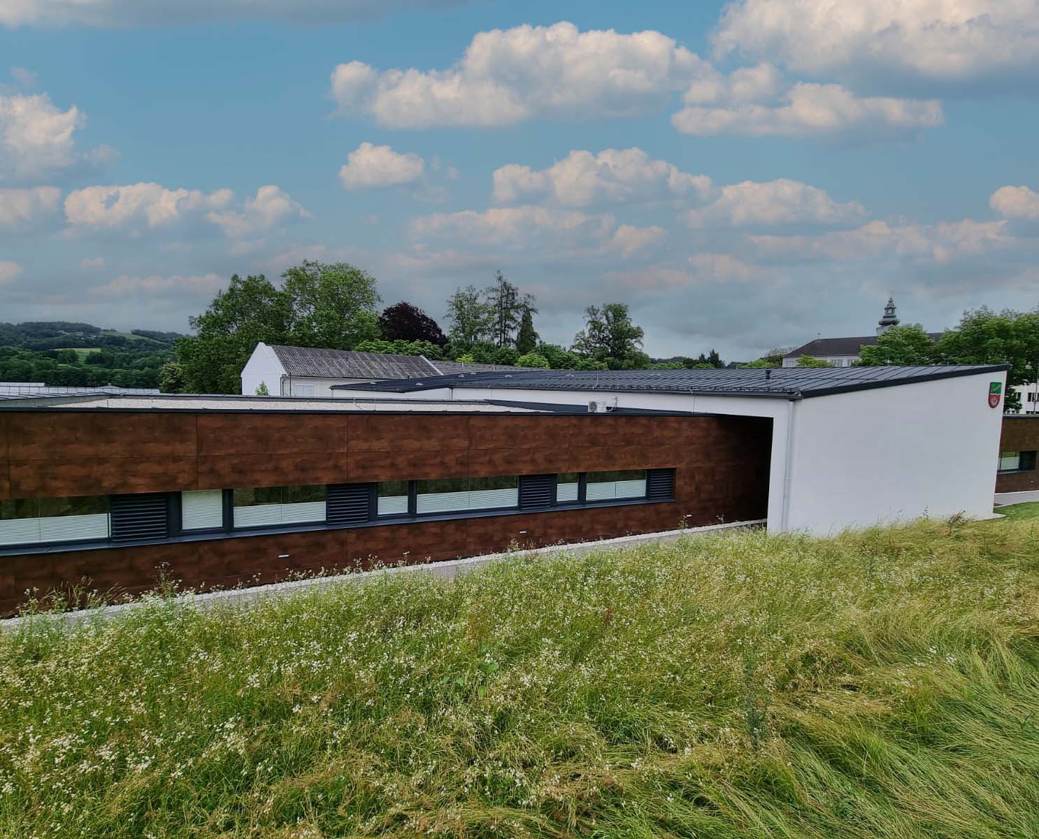 Modernes Gebäude mit einer Fassade aus braunem Holz und weißen Wänden, umgeben von einer Blumenwiese, unter blauem Himmel mit einigen Wolken.