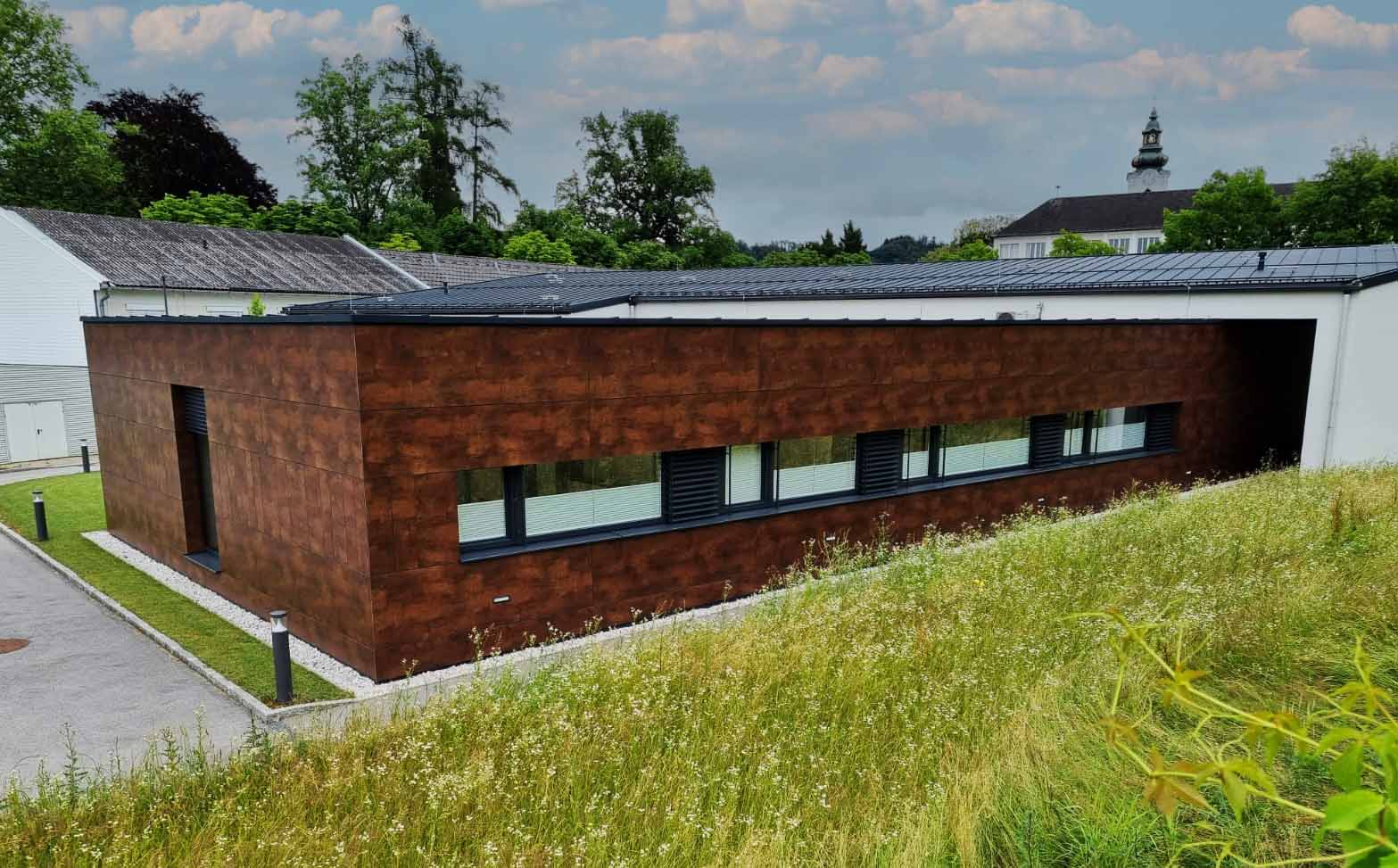 Modernes Gebäude mit rostfarbenen Fassadenplatten und langer Fensterfront, umgeben von Gras und Bäumen.