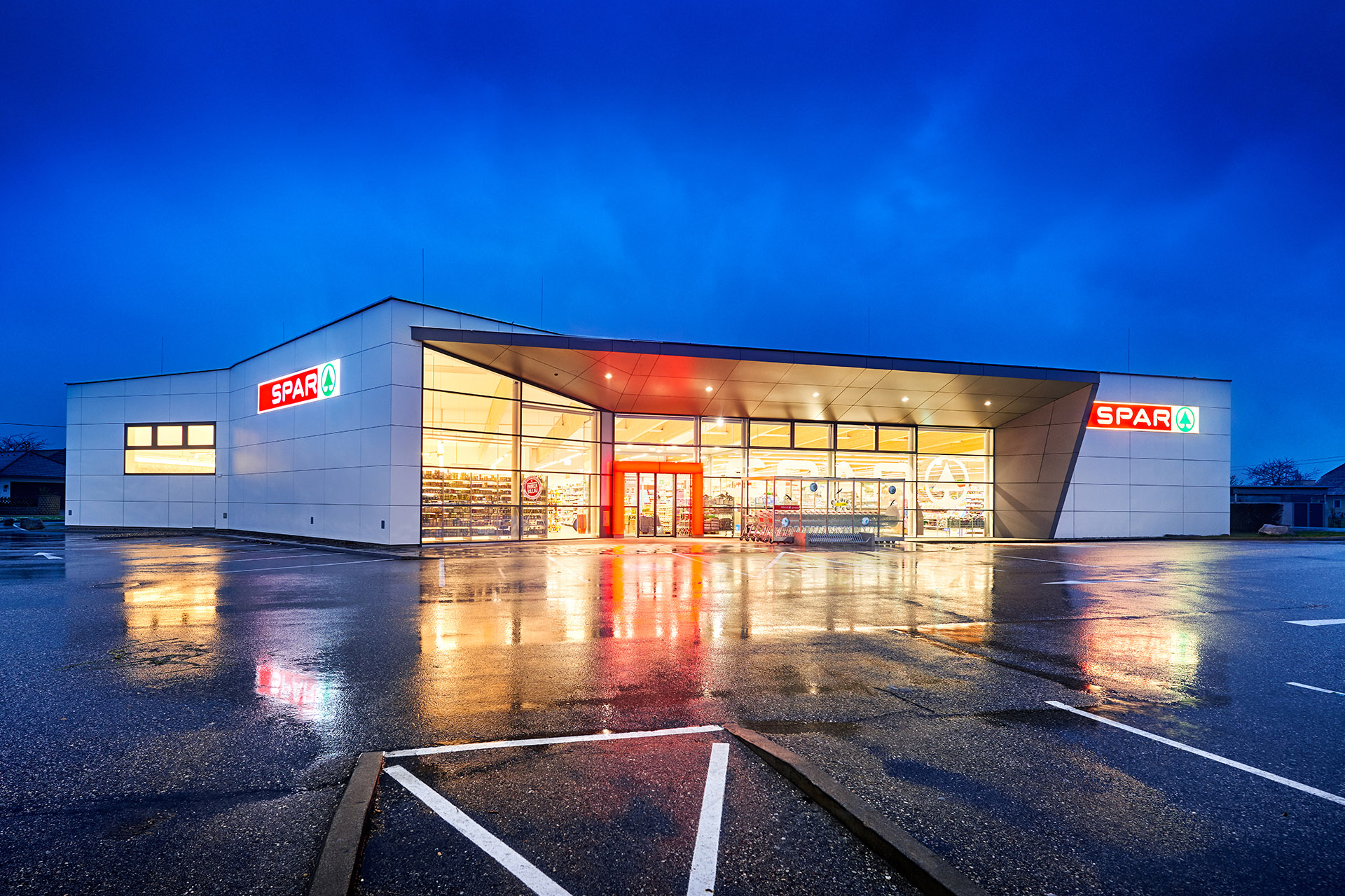 Modernes Spar-Einkaufszentrum bei Abenddämmerung mit leuchtendem Eingang und Reflexionen auf nasser Parkfläche.