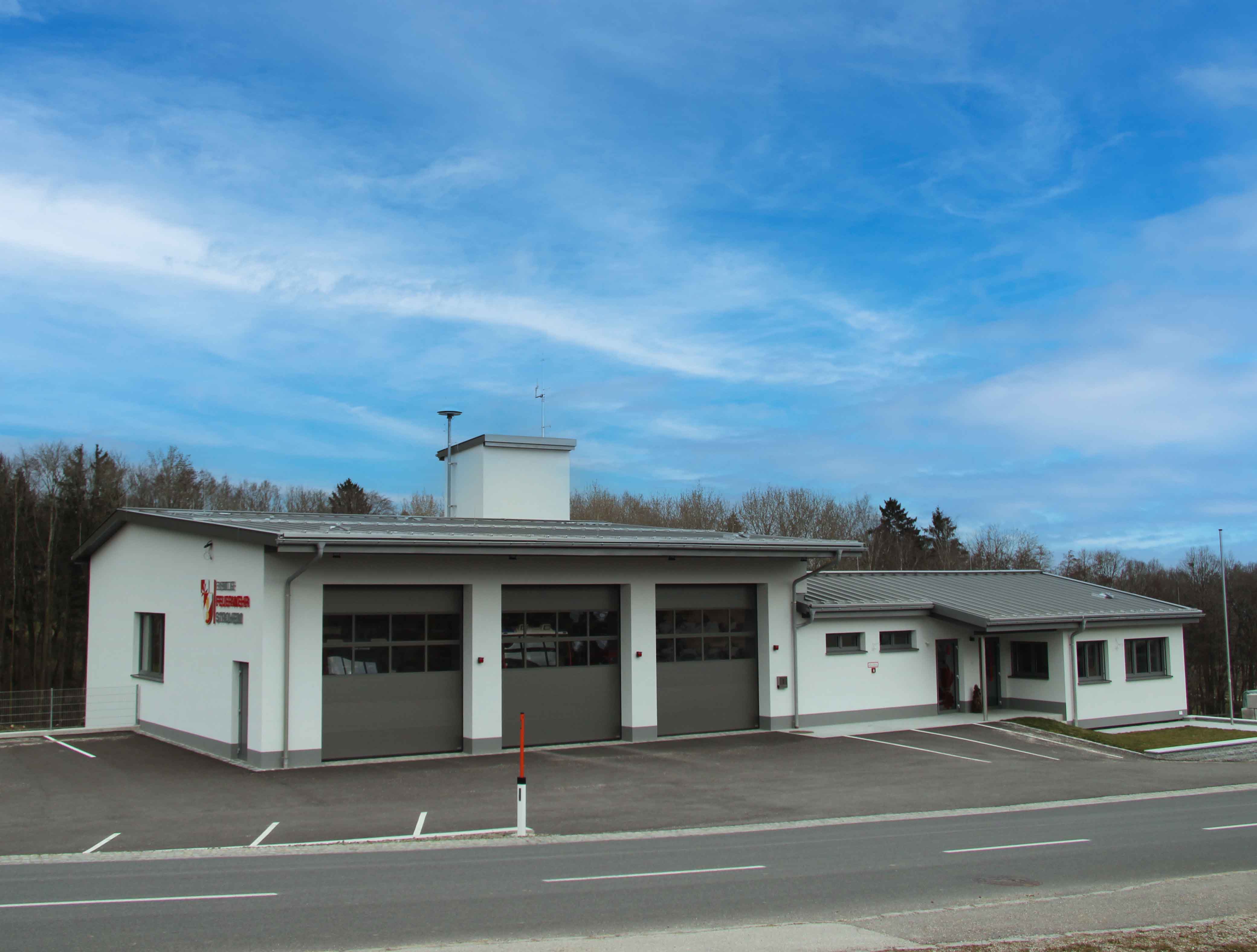 Modernes Feuerwehrhaus mit drei großen Garagentoren und Parkplatz, vor blau bewölktem Himmel.