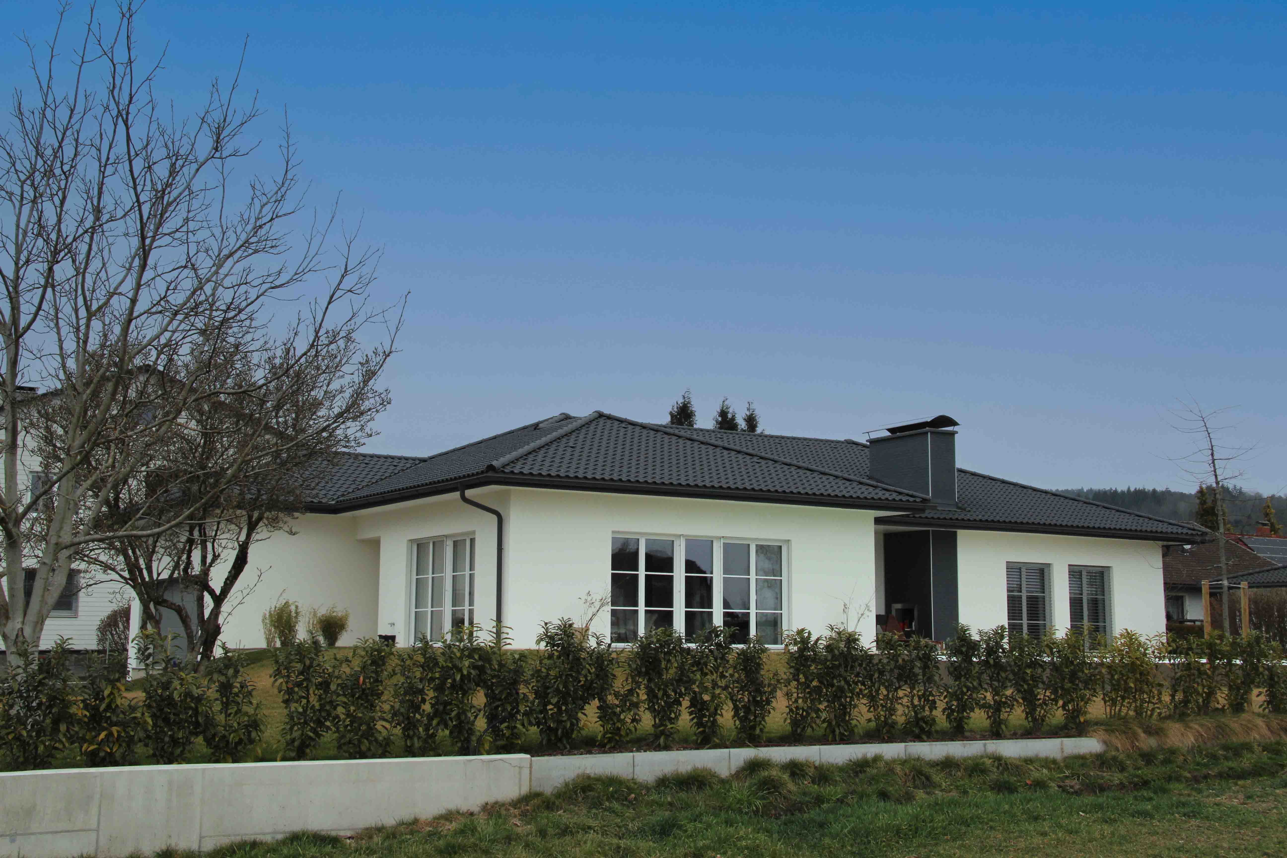 Ein modernes, einstöckiges Haus mit weißer Fassade, dunklem Satteldach und großen Fenstern, umgeben von einem Garten mit Hecken und kahlen Bäumen.