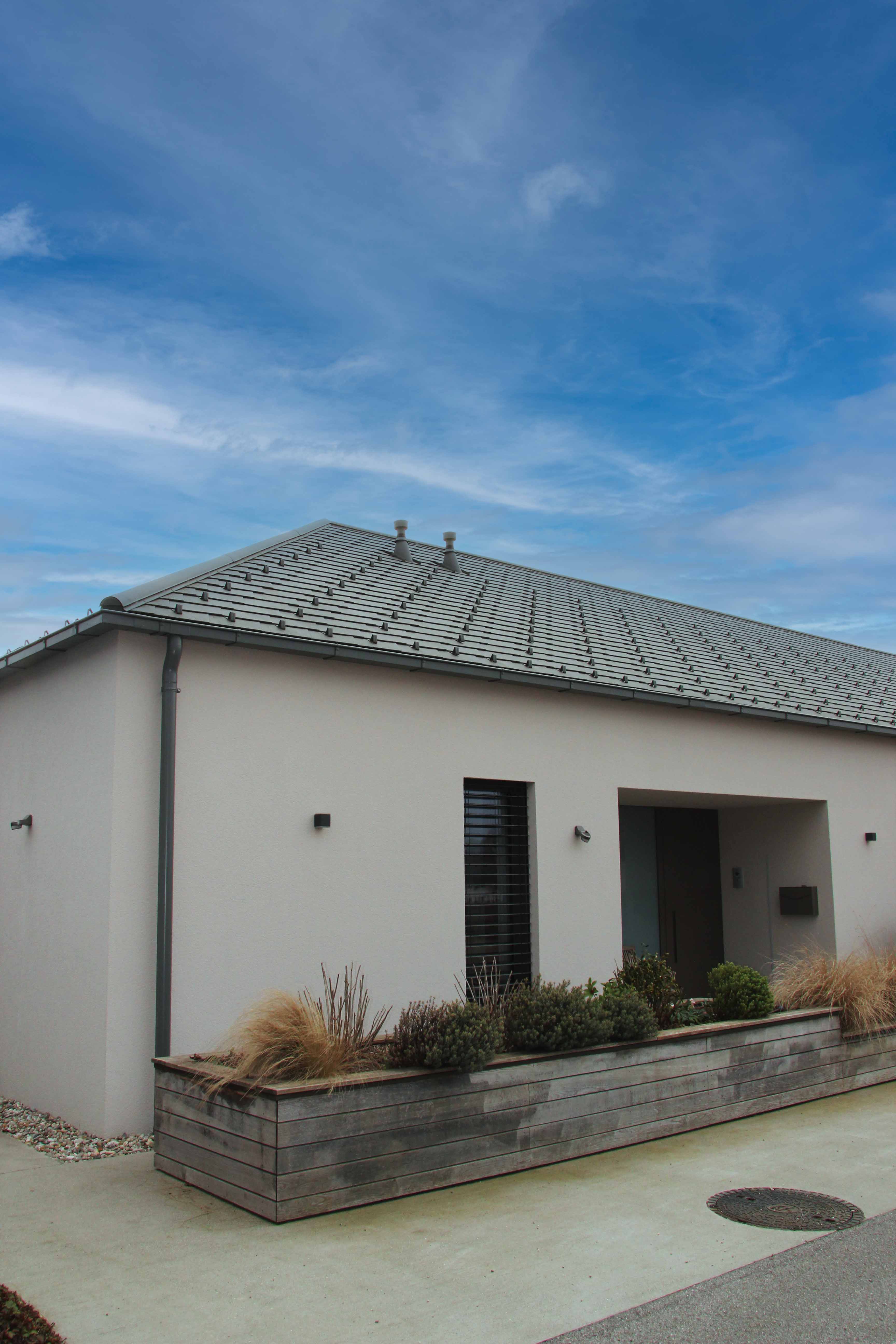 Modernes Einfamilienhaus mit grauem Satteldach, weißen Wänden und einem großen Holzpflanzkasten mit Sträuchern und Gras vor dem Eingang.
