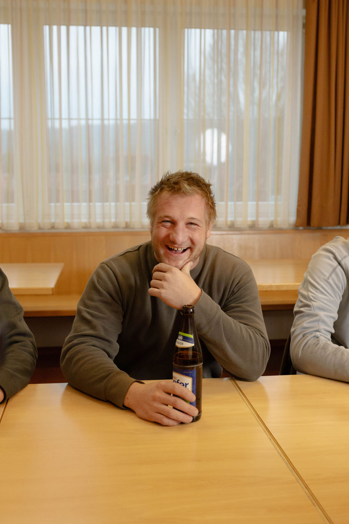 Mann mit braunem Pullover sitzt an einem Holztisch und hält eine Bierflasche, er lächelt in die Kamera.