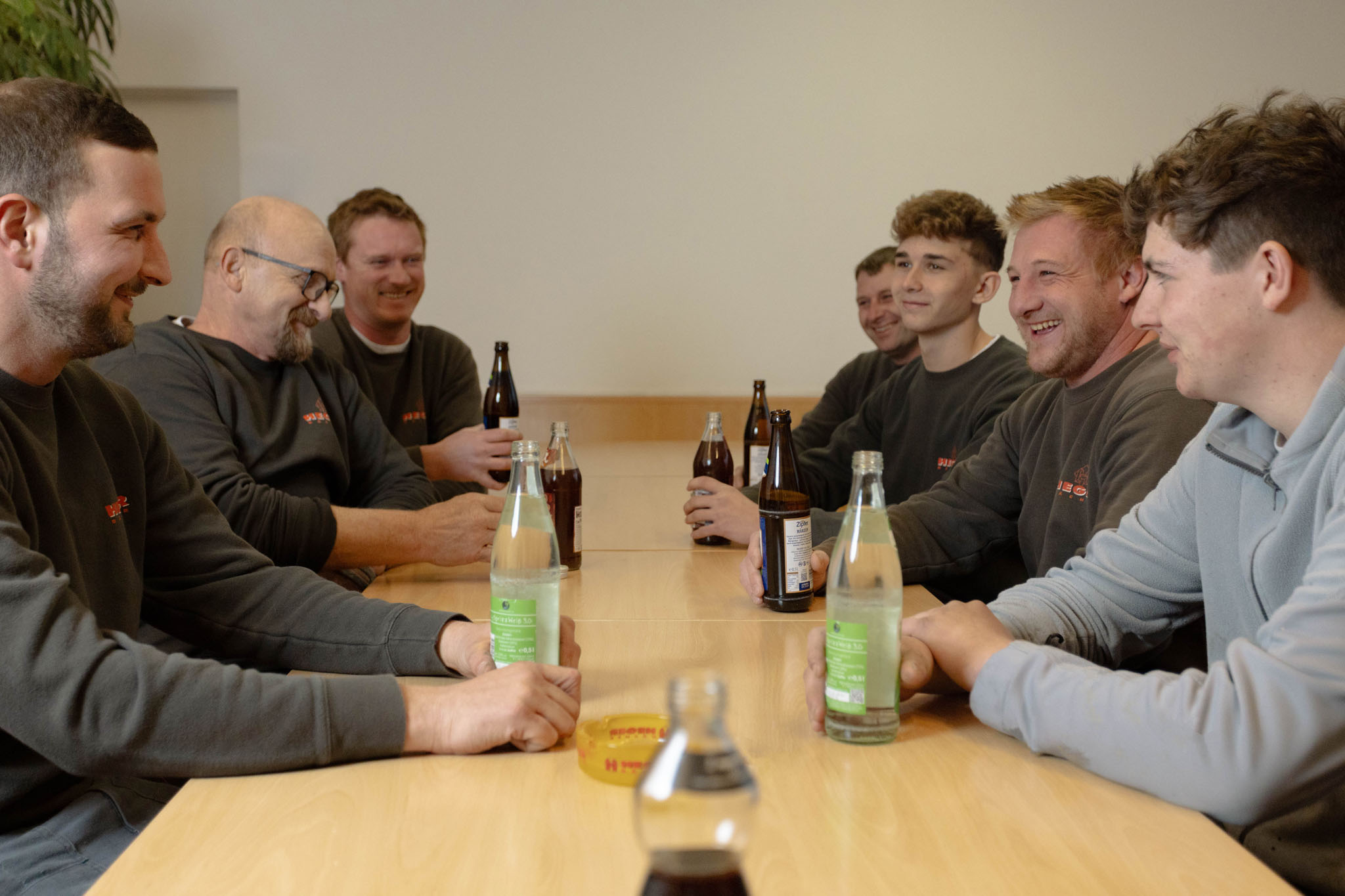 Acht Männer sitzen an einem Holztisch, trinken Flaschenbier und Mineralwasser und unterhalten sich.