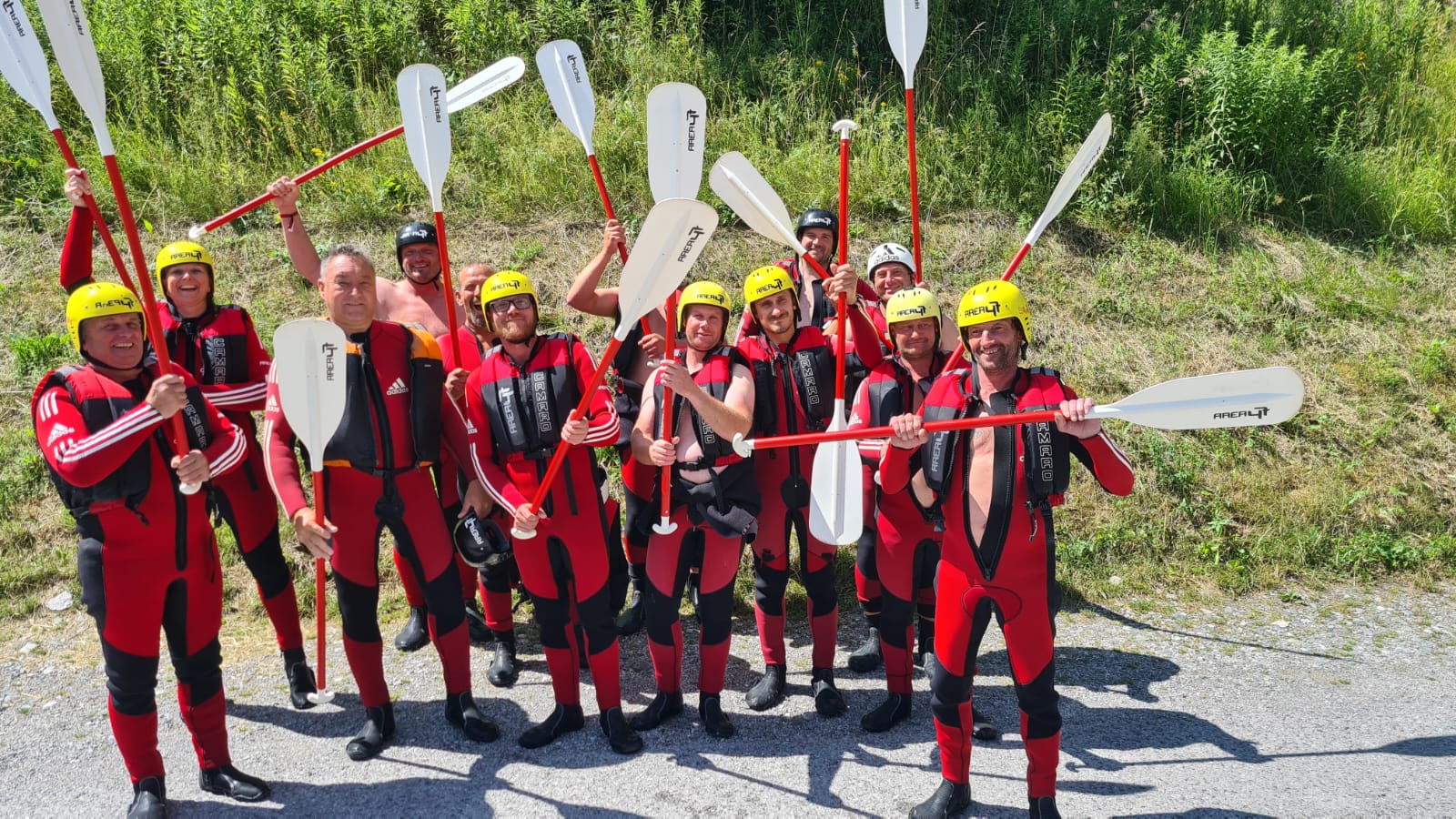 Gruppe von Erwachsenen in roten Neoprenanzügen und gelben Helmen, die Paddel hochhalten und draußen vor Gras stehen.