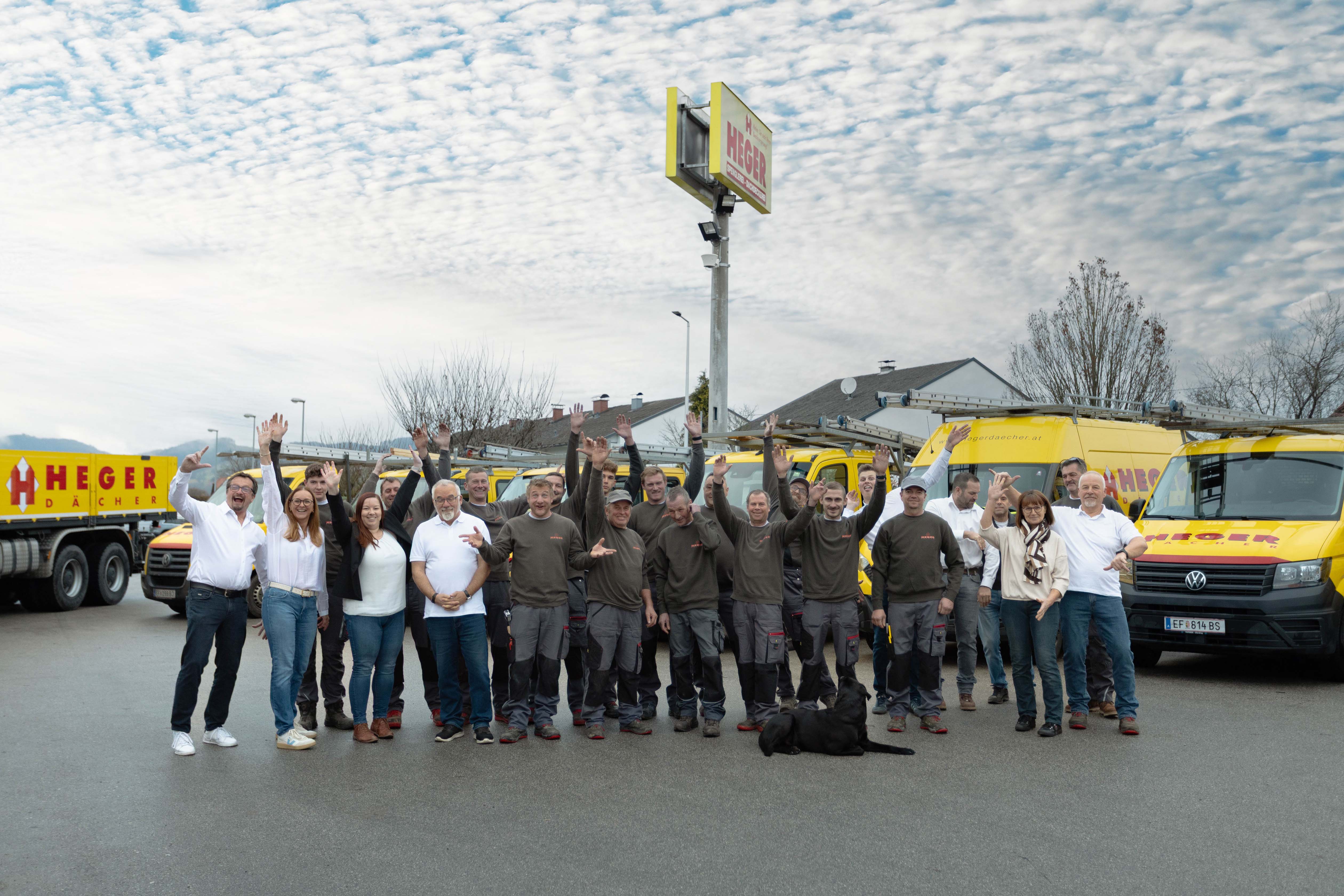 Gruppe von etwa zwanzig Personen, einige in Arbeitskleidung, die vor gelben Firmenfahrzeugen mit dem Schriftzug Heger Dächern stehen und in die Kamera winken, mit einem schwarzen Hund auf dem Boden.