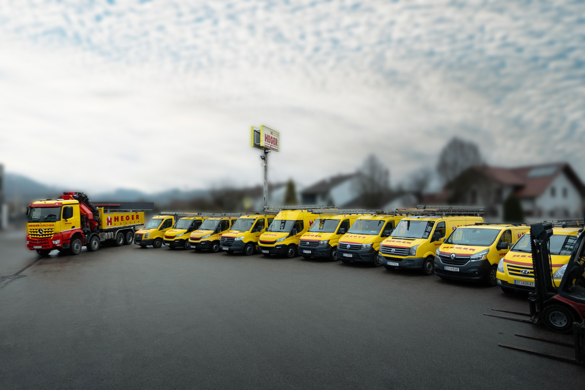 Gelbe Firmenfahrzeuge der Marke Heger geparkt in einer Reihe vor einem Firmenschild, darunter mehrere Transporter und ein großer LKW mit Kran.
