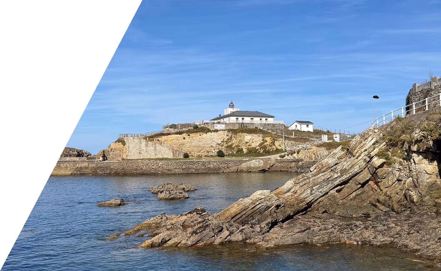 Rocky coastline with a lighthouse and buildings atop a cliff under a clear blue sky.