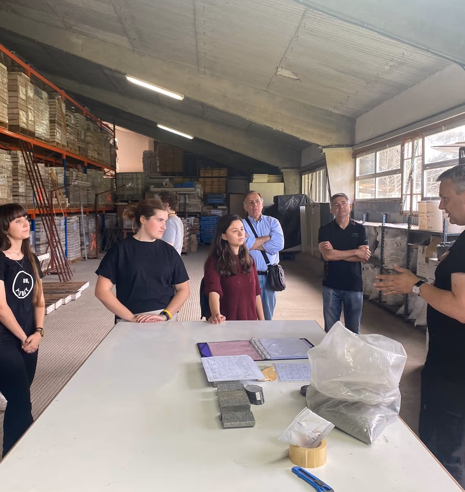 Group of people standing around a large table in a warehouse, listening to a man explaining while materials and documents are on the table.