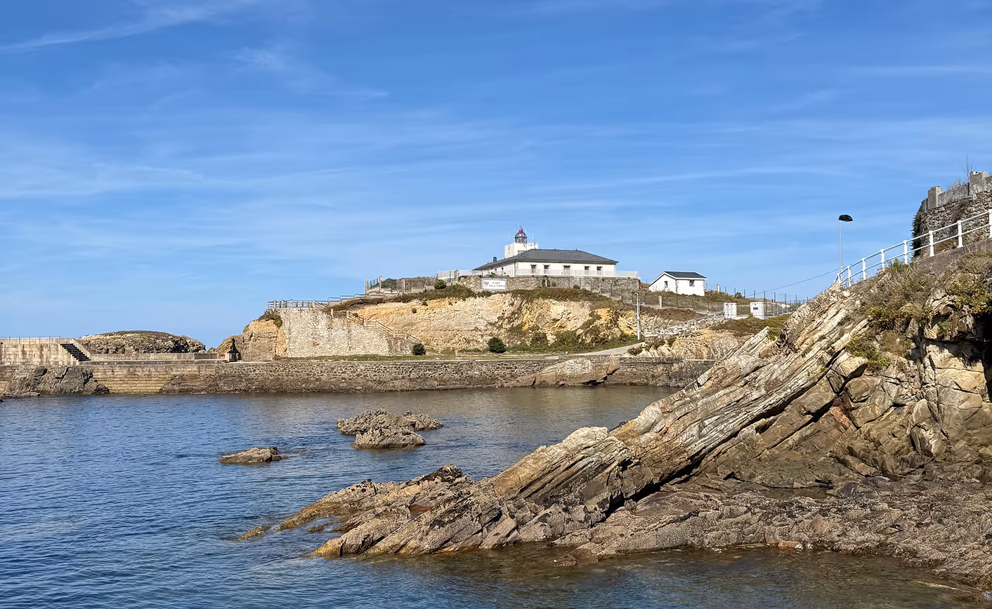 Rocky coastline with a lighthouse and buildings on a cliff under a clear blue sky.