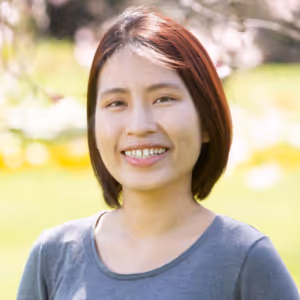Smiling woman with short reddish-brown hair wearing a gray top outdoors with a blurred natural background.