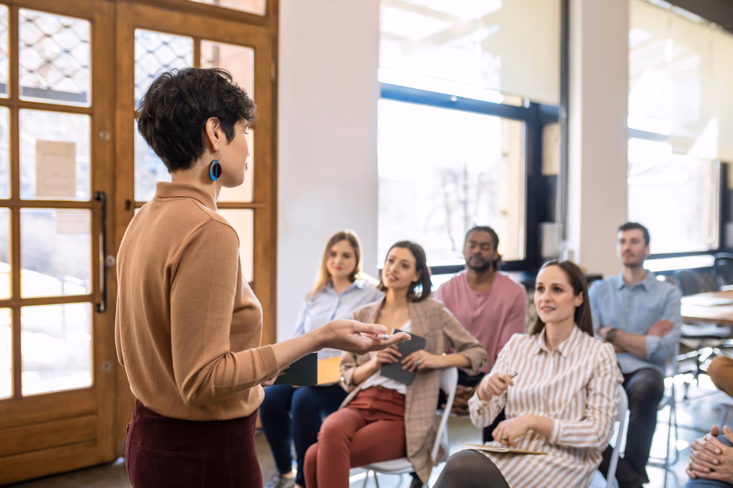 Woman in brown sweater leading a group discussion with attentive participants seated in a bright room.