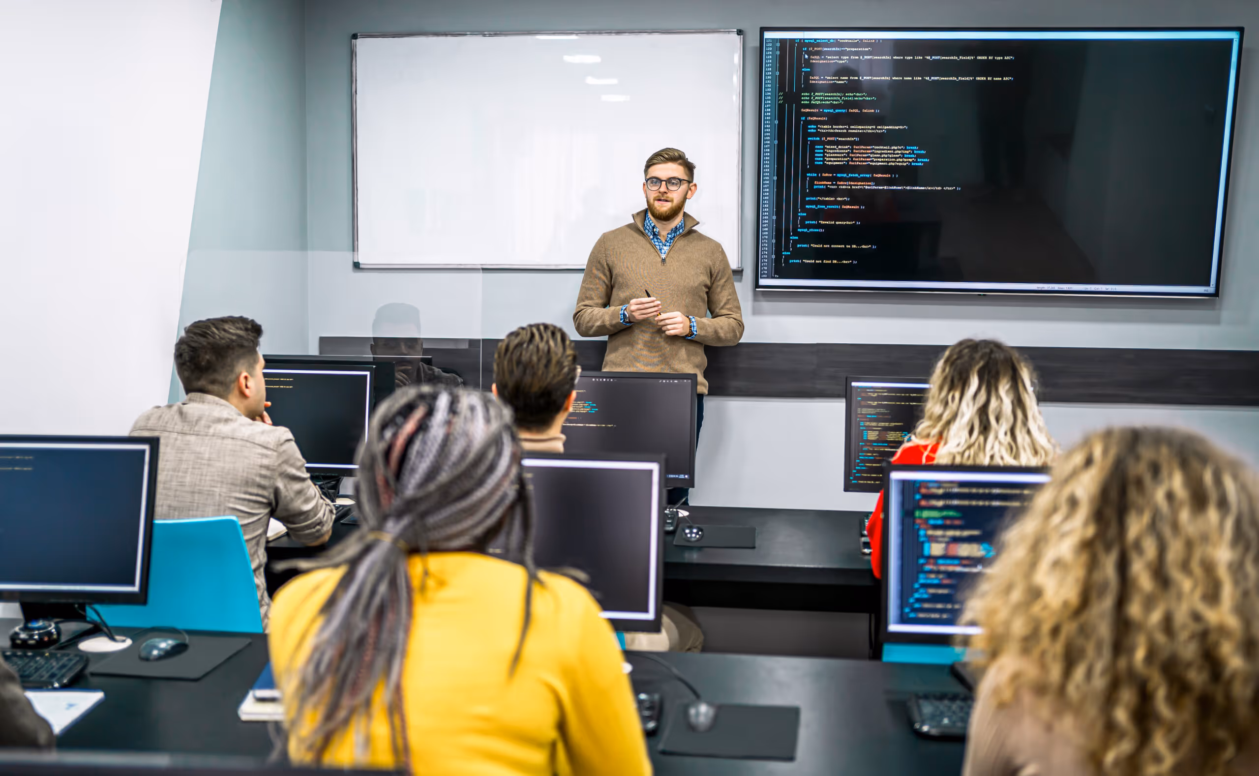 Instructor standing in front of a classroom teaching programming code displayed on a large screen to students at computer desks.