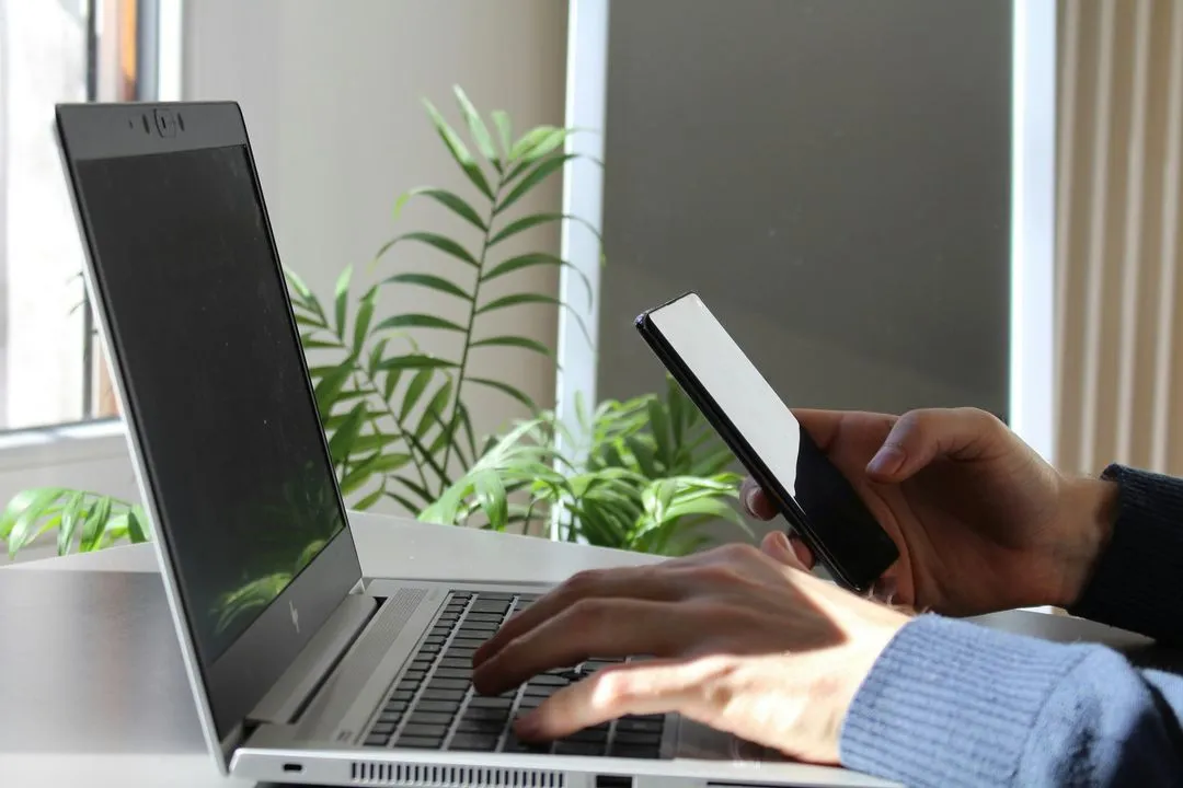 Hands holding a smartphone and typing on a laptop in a brightly lit room with green plants. The scene conveys multitasking or technology use.