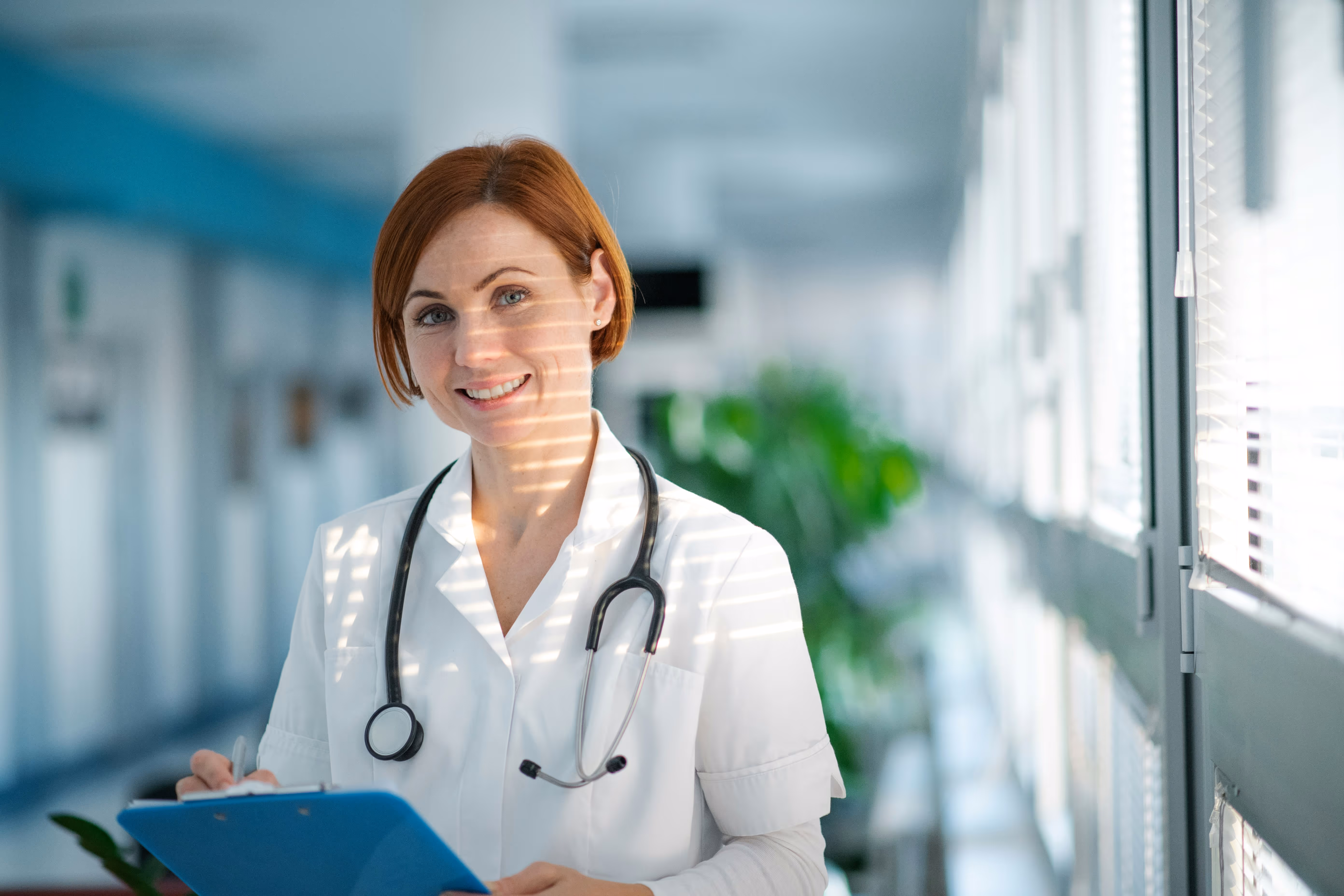 Female doctor holding a clipboard in a hospital corridor, representing patient care, medical staffing, or licensed healthcare professionals in clinical environments.